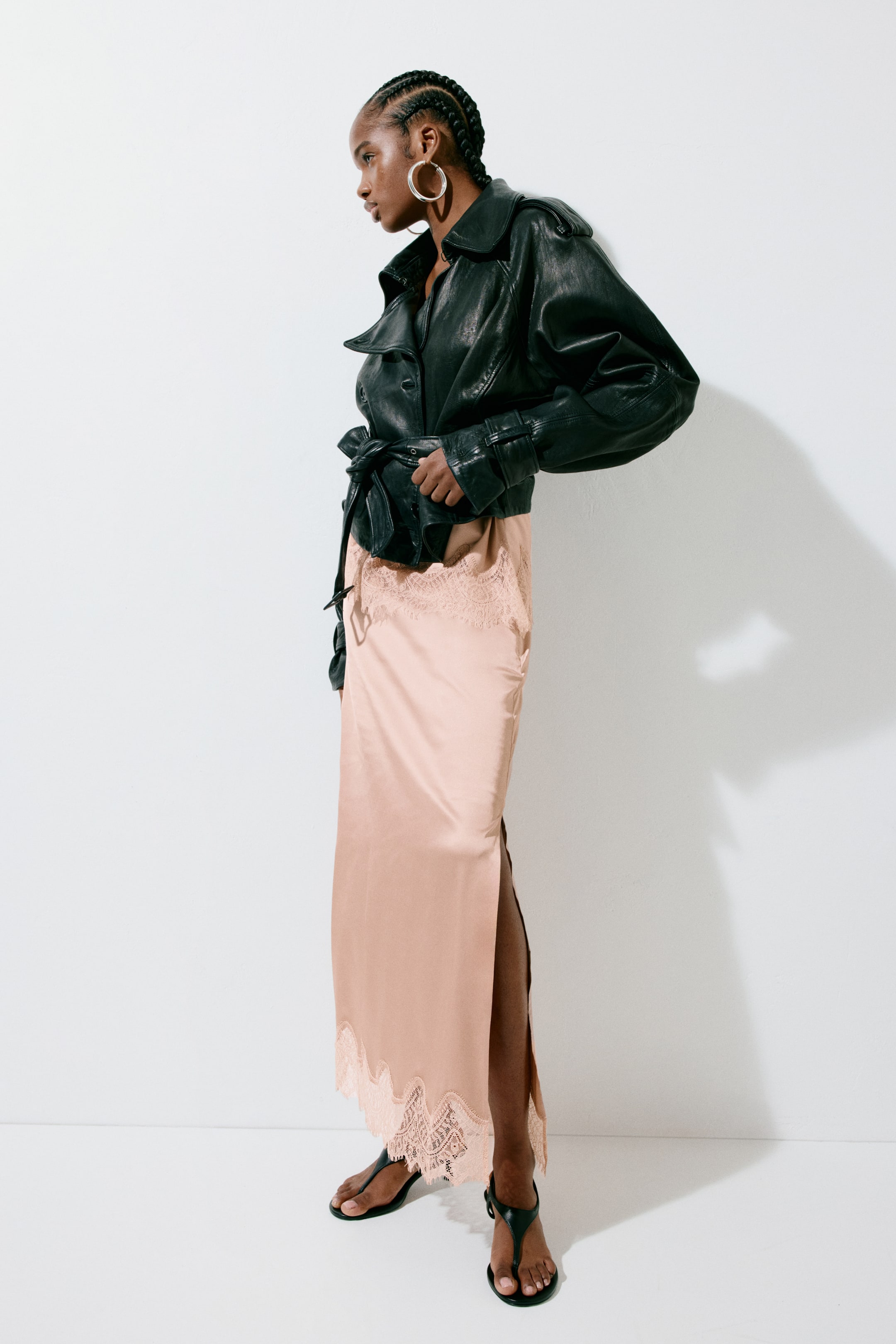 View larger image: A black leather jacket showcasing wide, pointed lapels, shoulder flaps, and a belted waist tied in a knot at the front. The cropped jacket features long sleeves with strapped cuffs and is viewed from a side-front angle.