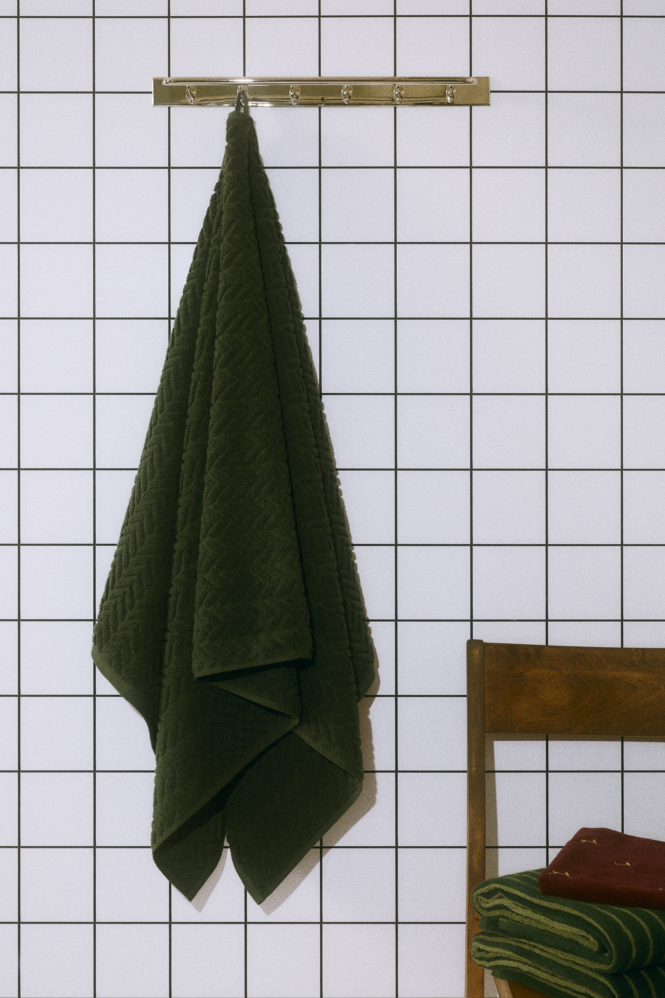 Een donkergroene badhanddoek van katoenbadstof hangt aan een kleine zwarte haak en toont een gestructureerd kabelbreipatroon in chevronstijl. De handdoek hangt verticaal en laat de zachte geweven textuur en afgewerkte randen zien.