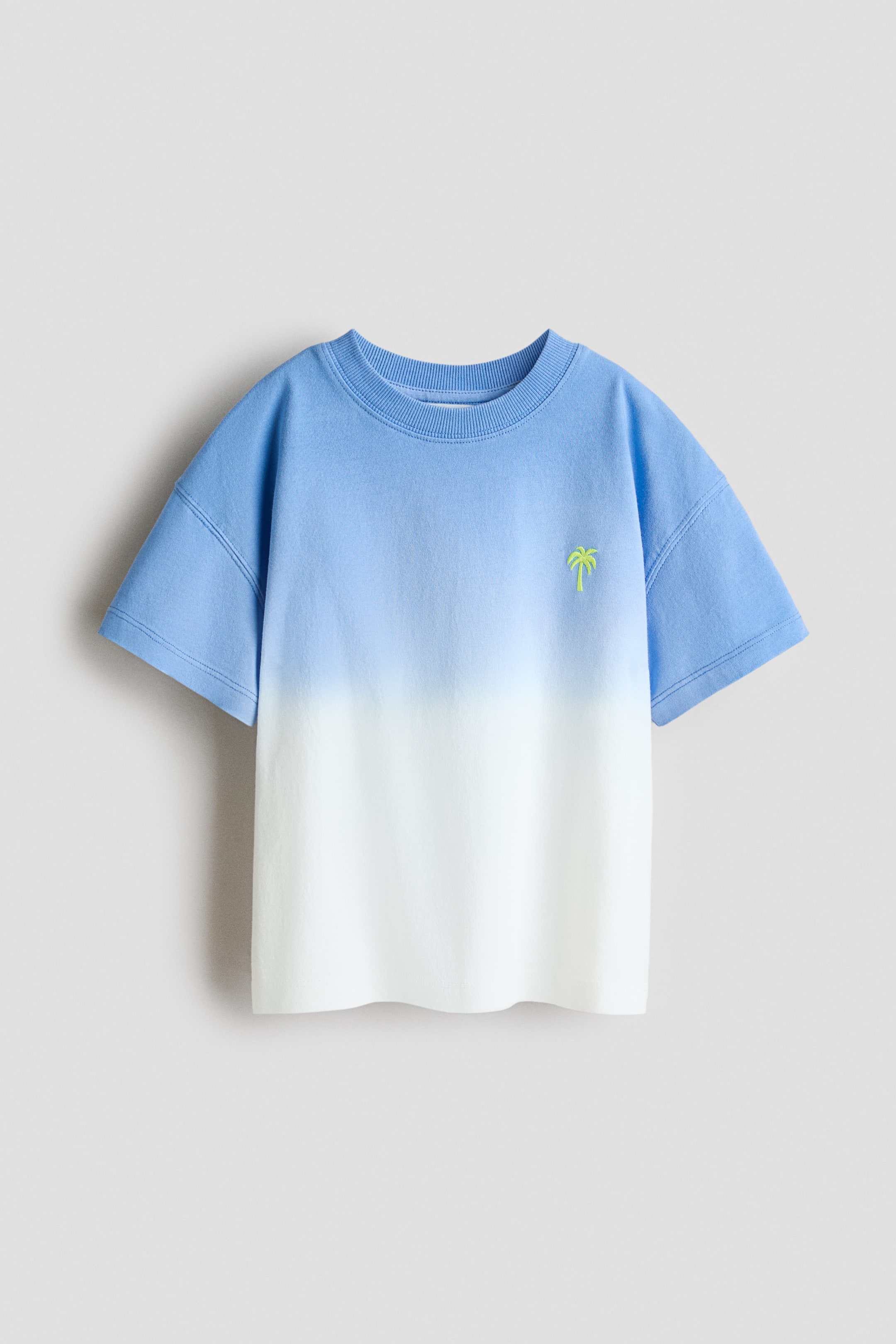拡大画像を表示: オーバーサイズのTシャツは、フロントにライトブルーからホワイトへのグラデーションデザインを施しています。リブ付きのクルーネック、ドロップショルダー、ストレートヘムが特徴です。左胸には小さなネオングリーンのヤシの木が刺繍されています。