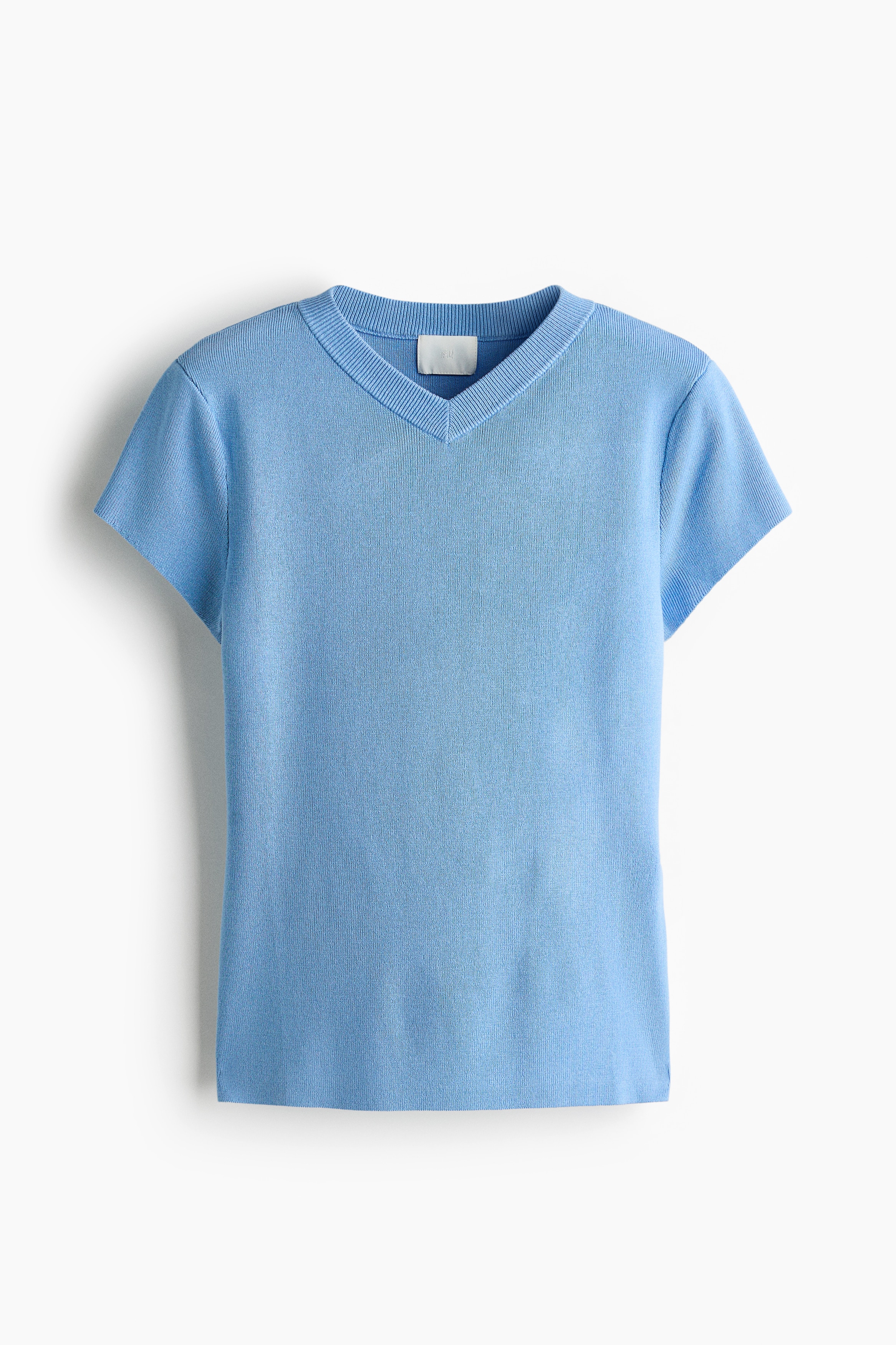 La modelo lleva puesta una playera ajustada de tejido fino en color azul claro, con cuello en V con ribete acanalado y mangas cortas tipo cap. La modelo combina la playera con jeans de mezclilla azules, con las manos en los bolsillos delanteros.