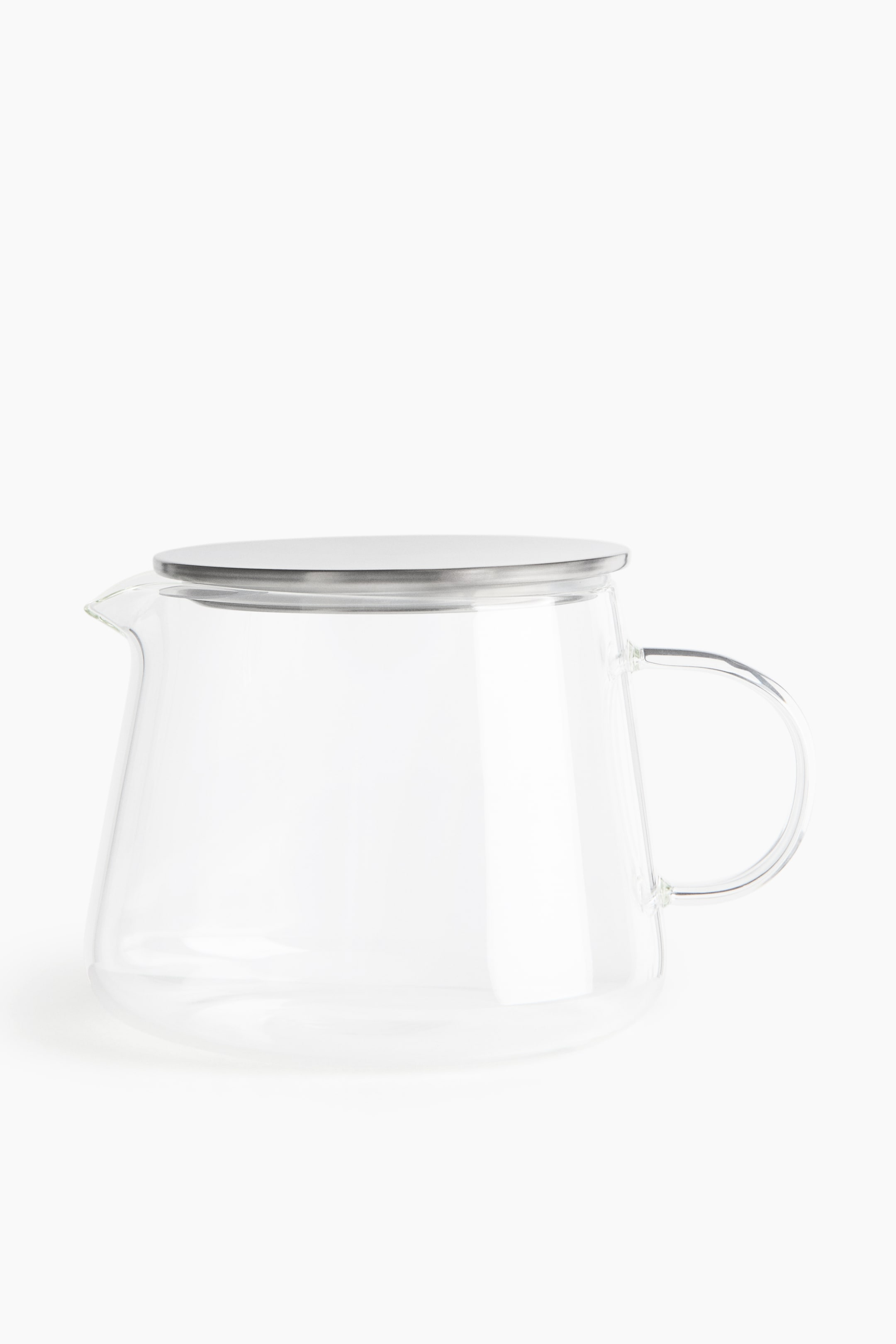 Voir une image plus grande: Une théière en verre transparent est présentée de profil, avec un corps large et effilé, un petit bec verseur d’un côté et une poignée incurvée en verre de l’autre. Elle est surmontée d’un couvercle plat et circulaire en acier inoxydable.