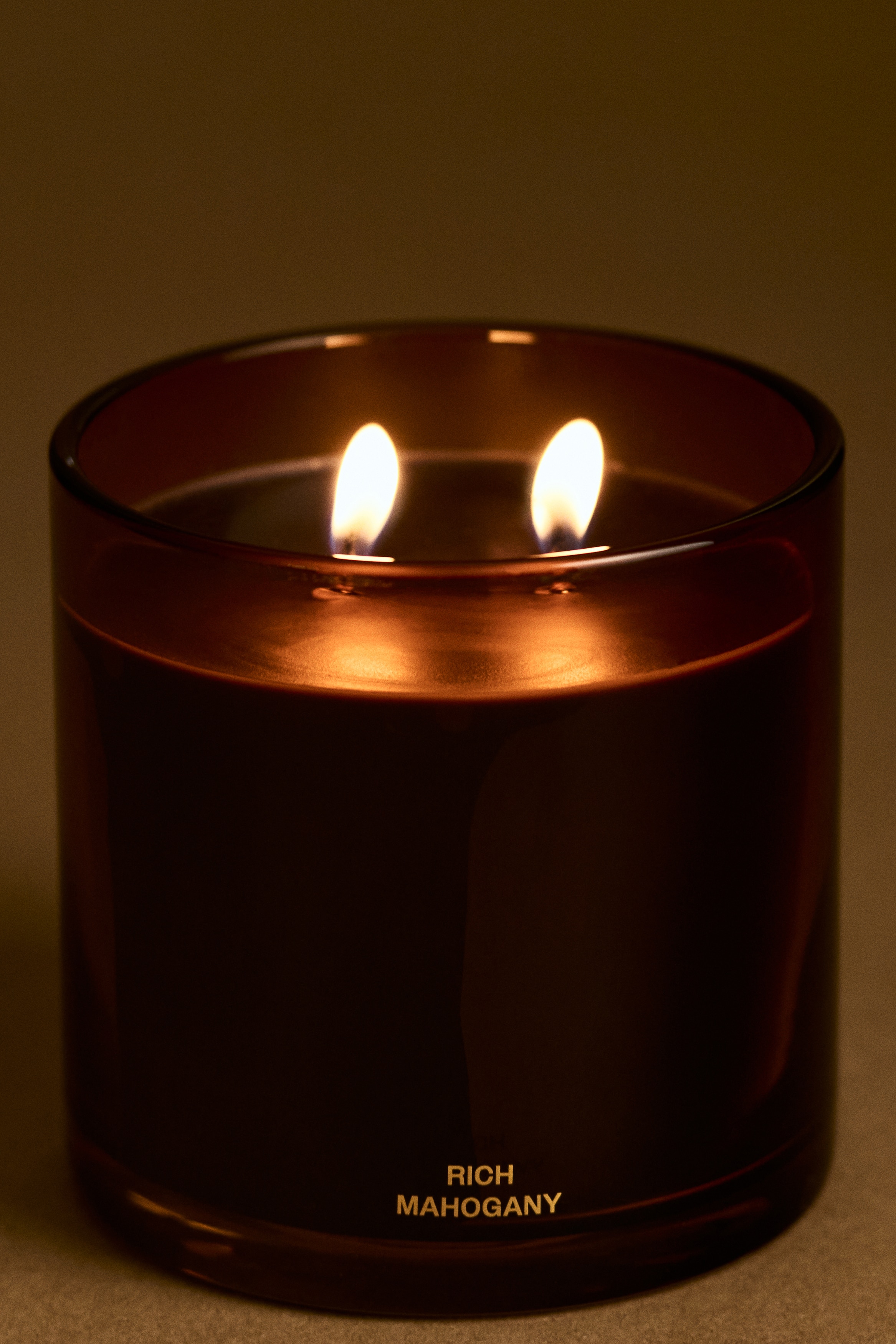A scented candle in a cylindrical amber glass vessel is positioned facing forward, featuring dark wax with two visible wicks. The lower front of the glass container displays the words "RICH MAHOGANY" in small gold capital letters above a thick, clear base.