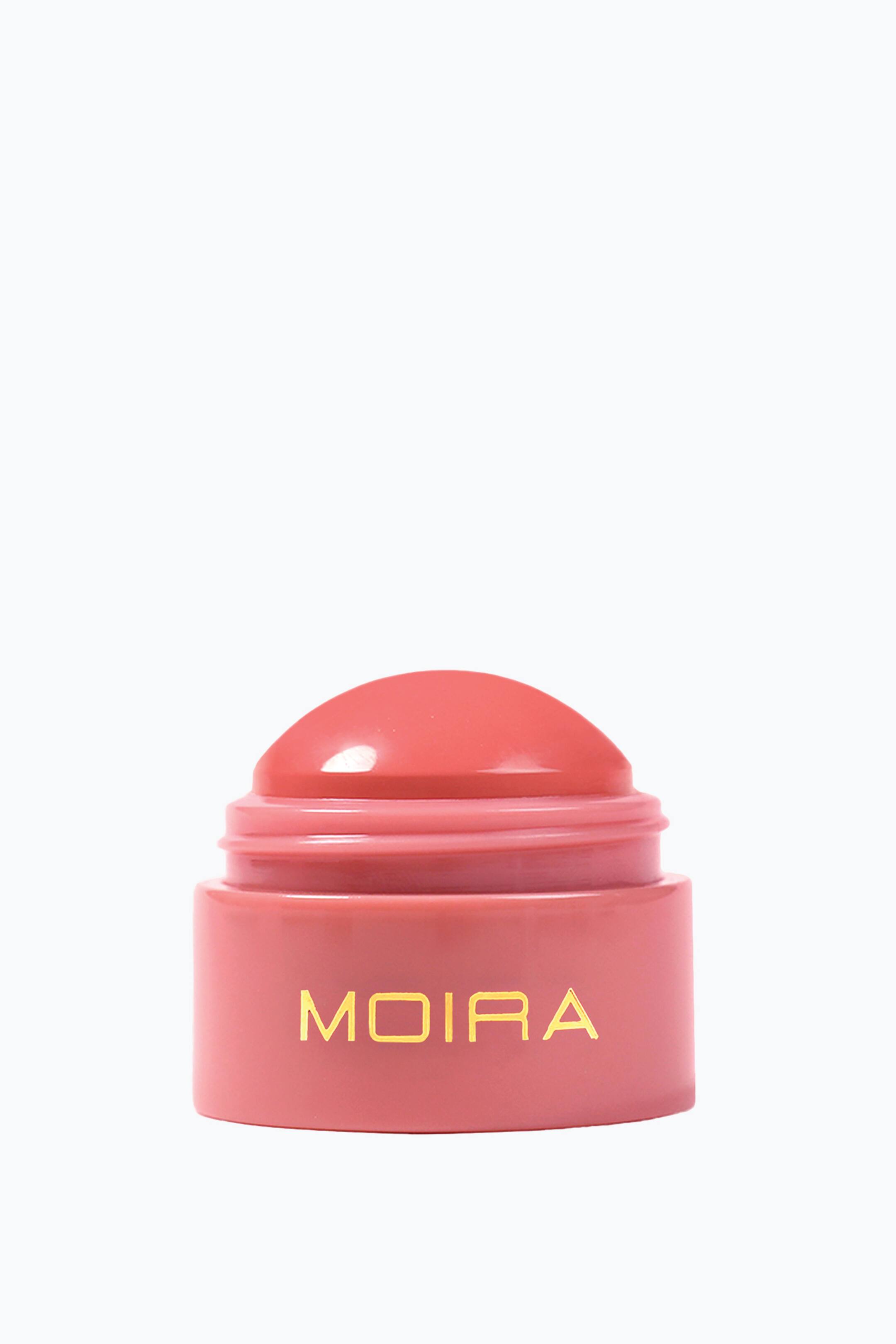 Se større billede: En cremeblush-balm står oprejst og fremviser et glat, kuppelformet koralrosa produkt, der stikker op fra en matchende lyserød cylindrisk base. Forsiden af beholderen viser brandnavnet "MOIRA" i gyldne, versaler.
