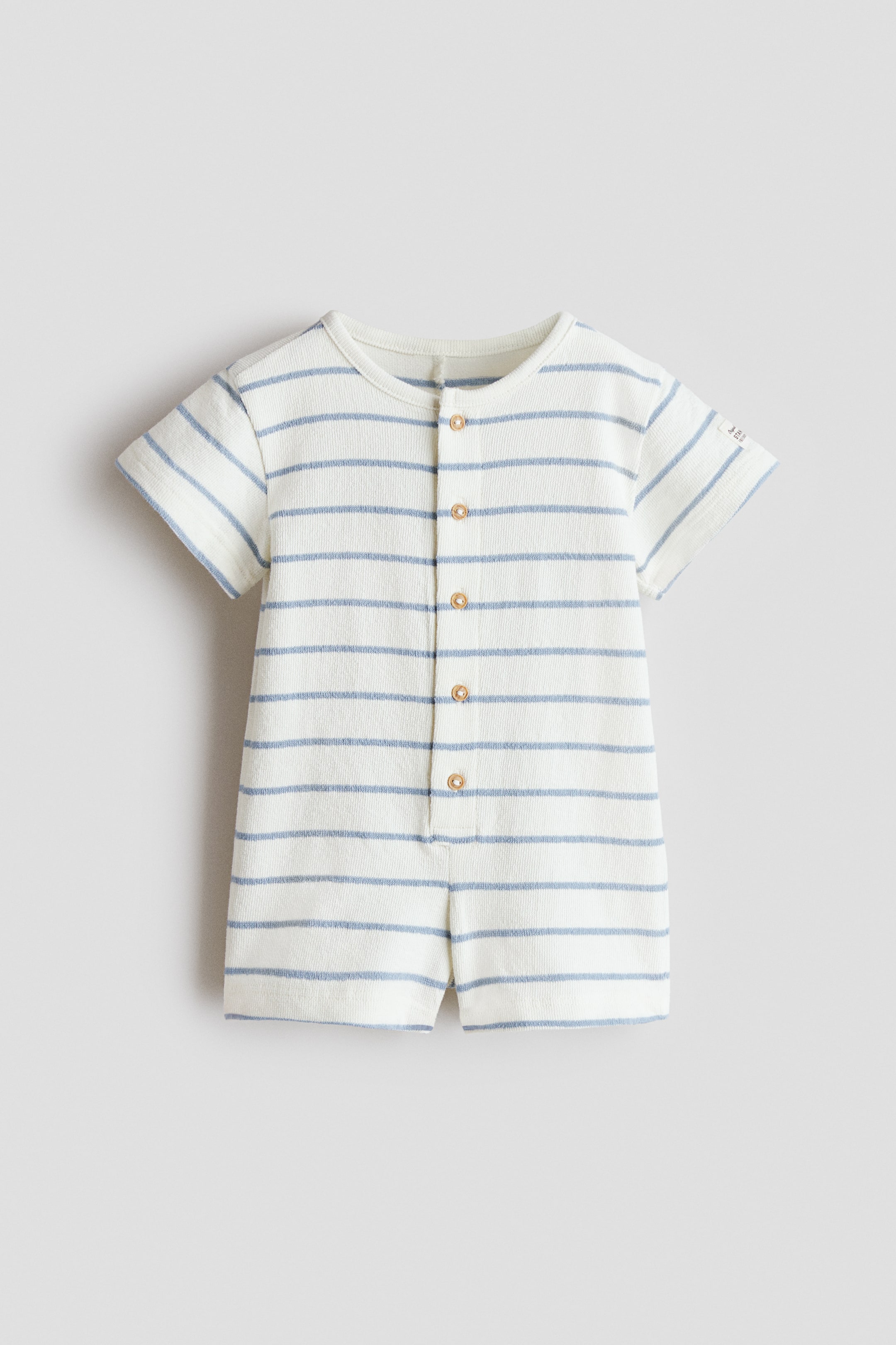 View larger image: An infant romper is showcasing a textured cream fabric with horizontal blue stripes, featuring a round neckline, short sleeves, and a front placket with six light brown buttons.
