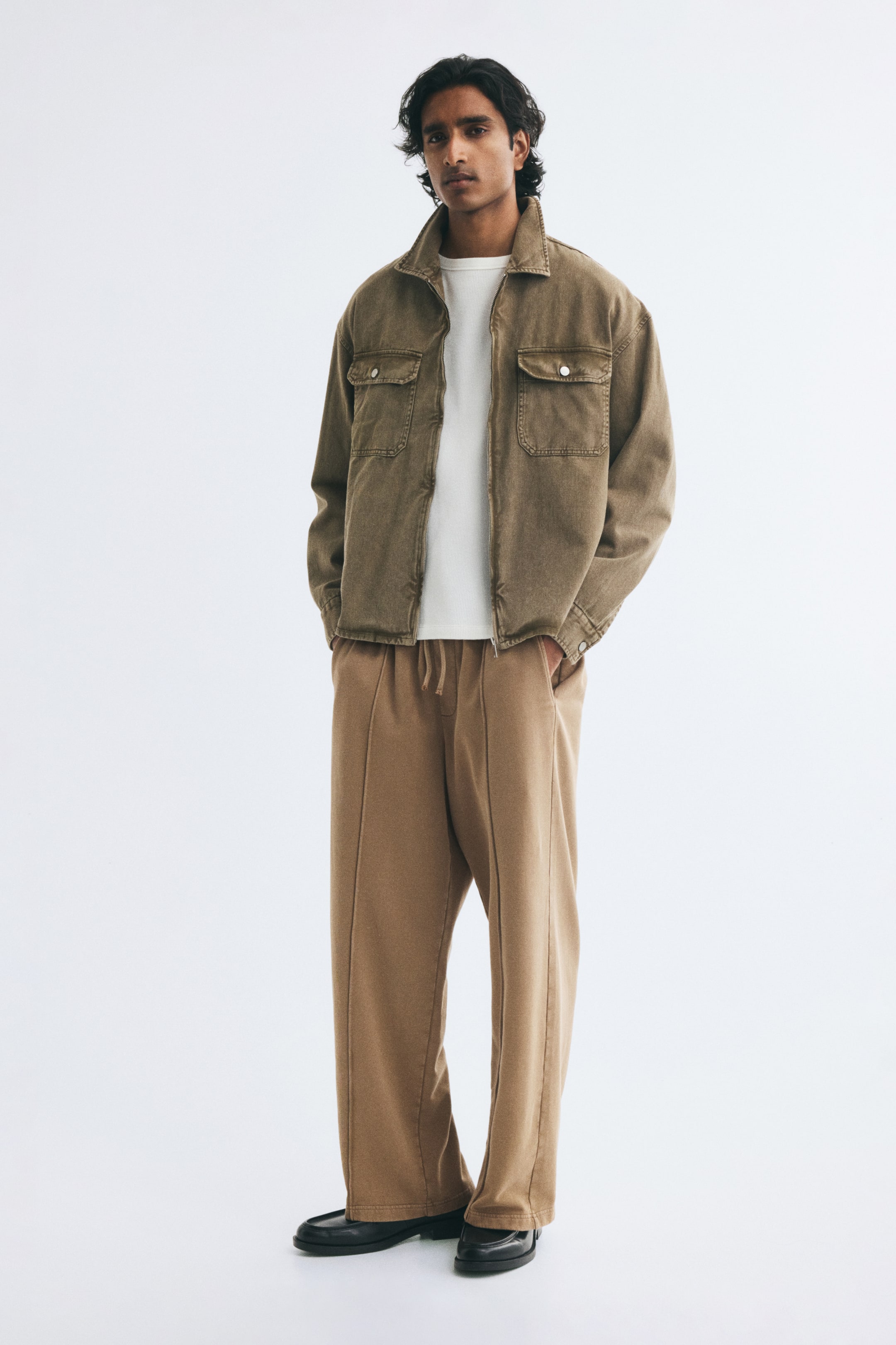View larger image: The model is wearing an olive-brown denim jacket with a collar and chest flap pockets, worn open over a white T-shirt. The jacket is paired with tan, wide-leg drawstring trousers, and the model is standing with hands in the pockets.