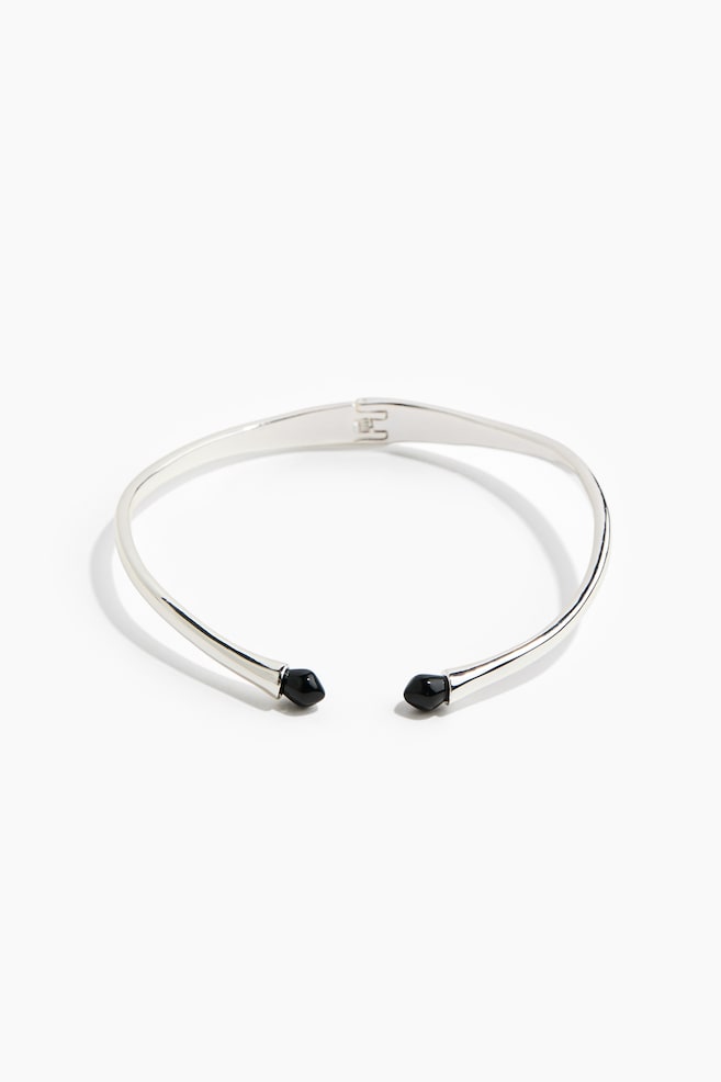 A silver-toned metal collar necklace is shown from the front, featuring a rigid, circular band with a small hinge at the back centre. The open ends at the front taper slightly and are capped with pointed, glossy black gemstone tips. - 1