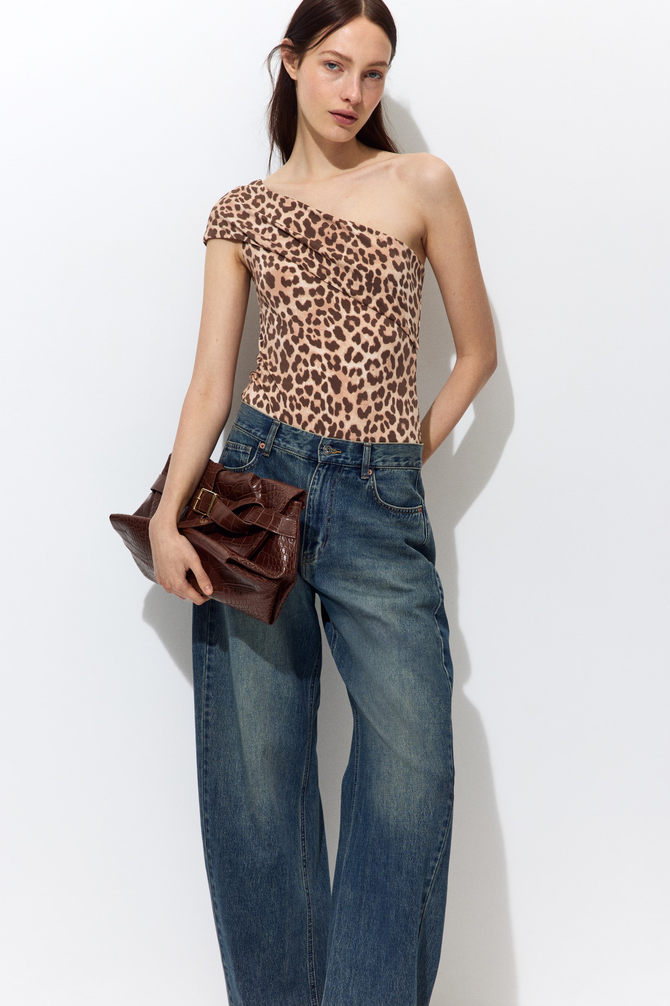 Ampliar la imagen: La modelo lleva puesto un body de un solo hombro con un pliegue en la parte superior, con un estampado de leopardo. El body se combina con jeans azules de pierna ancha, y la modelo sostiene un bolso marrón con grabado de cocodrilo.