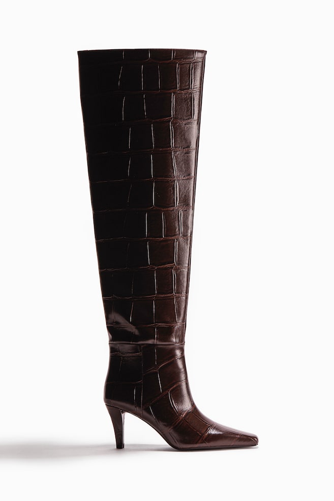 Ein dunkelbrauner Overknee-Stiefel aus krokodilgeprägtem beschichtetem Stoff steht und zeichnet sich durch eine spitze Zehenpartie und einen schlanken Kegelabsatz aus. - 1