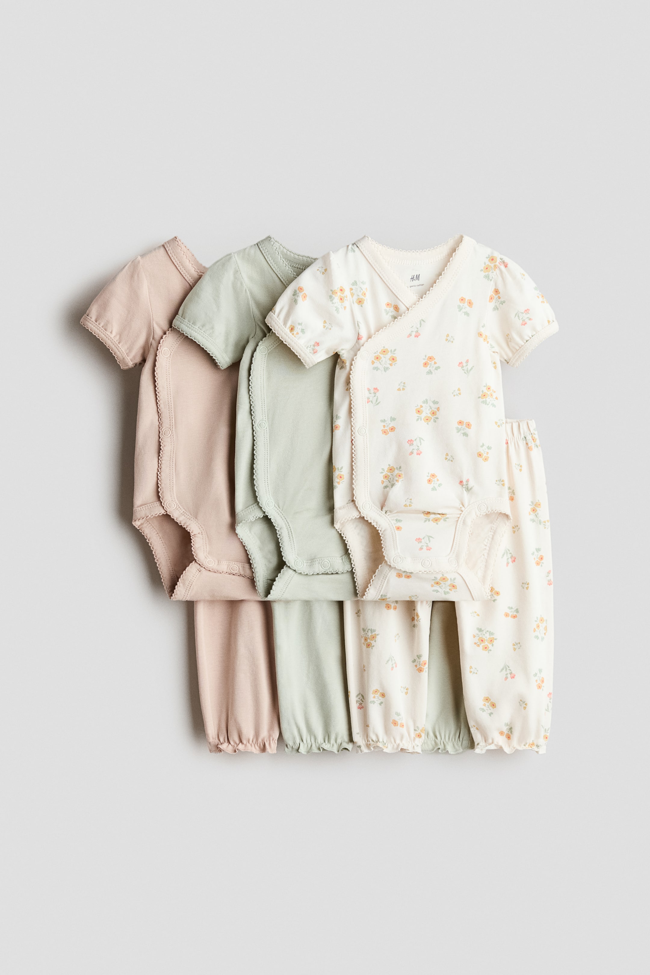 Grösseres Bild ansehen: Ein Set aus drei kurzärmeligen Baby-Bodys und passenden Hosen wird frontseitig präsentiert. Die Bodys in Altrosa, Salbeigrün und einem cremeweißen Blumenmuster verfügen über Wickelvorderseiten mit Picot-verzierten Kanten, Druckknöpfen an den Seiten und am Boden sowie gerafften Beinabschlüssen, während die dazugehörigen Hosen mit frill-verzierten, elastischen Säumen aufwarten.