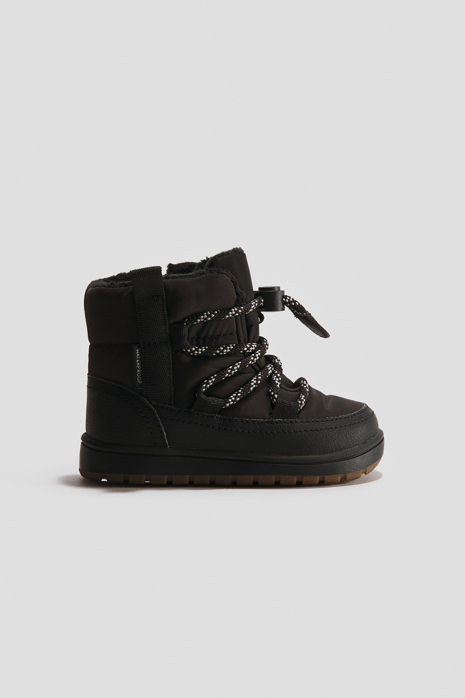 Ein schwarzer Winterstiefel ist in Seitenansicht nach rechts ausgerichtet und zeigt ein gestepptes Textilobermaterial mit strukturiertem Mudguard sowie kreuzweise verlaufende SchnÃ¼rsenkel, die mit einem Toggle-Verschluss gesichert sind. Der Stiefel prÃ¤sentiert ein schwarzes Fleecefutter, das am Schaftkragen hervorschaut, sowie eine strukturierte braune GummiauÃensohle.