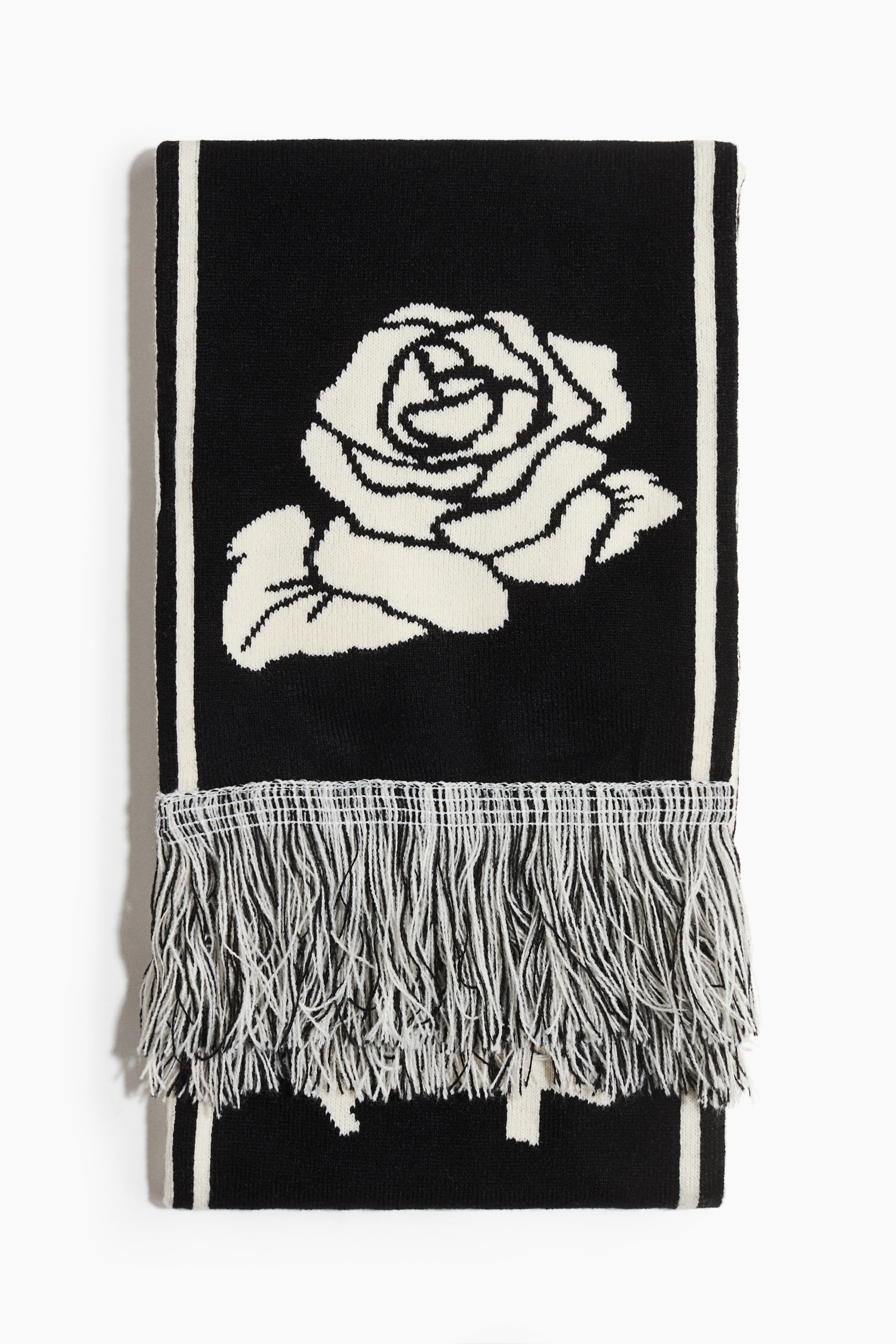 查看大圖: 一條圍巾平放著，展示其正面設計。它以一個大型的米白色玫瑰圖案為特色，位於黑色針織背景的中央，長邊沿著細米白色條紋作為邊框，並在一端配有黑色和米白色的流蘇作為收尾。