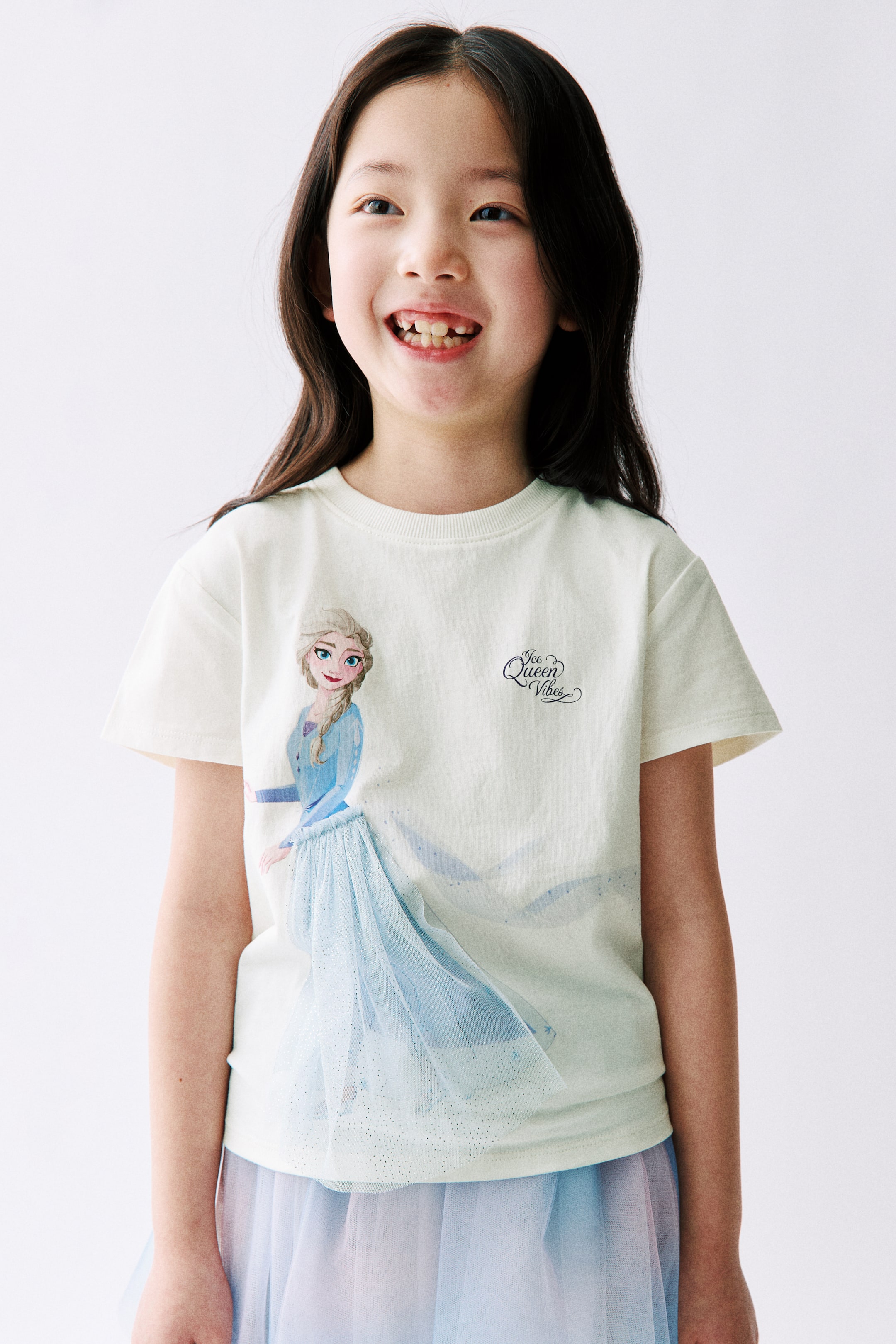 拡大画像を表示: クリーム色のTシャツに、左向きで立つ『アナと雪の女王』のエルサのグラフィックプリントが施されています。彼女のライトブルーのドレスのスカートは、レイヤードチュールの生地として広がっています。このチュール生地は、Tシャツの下に着用されたライトブルーとピンクのレイヤードチュールスカートと視覚的に一体化しています。右胸には「アイス・クイーン・バイブス」という文字もプリントされています。