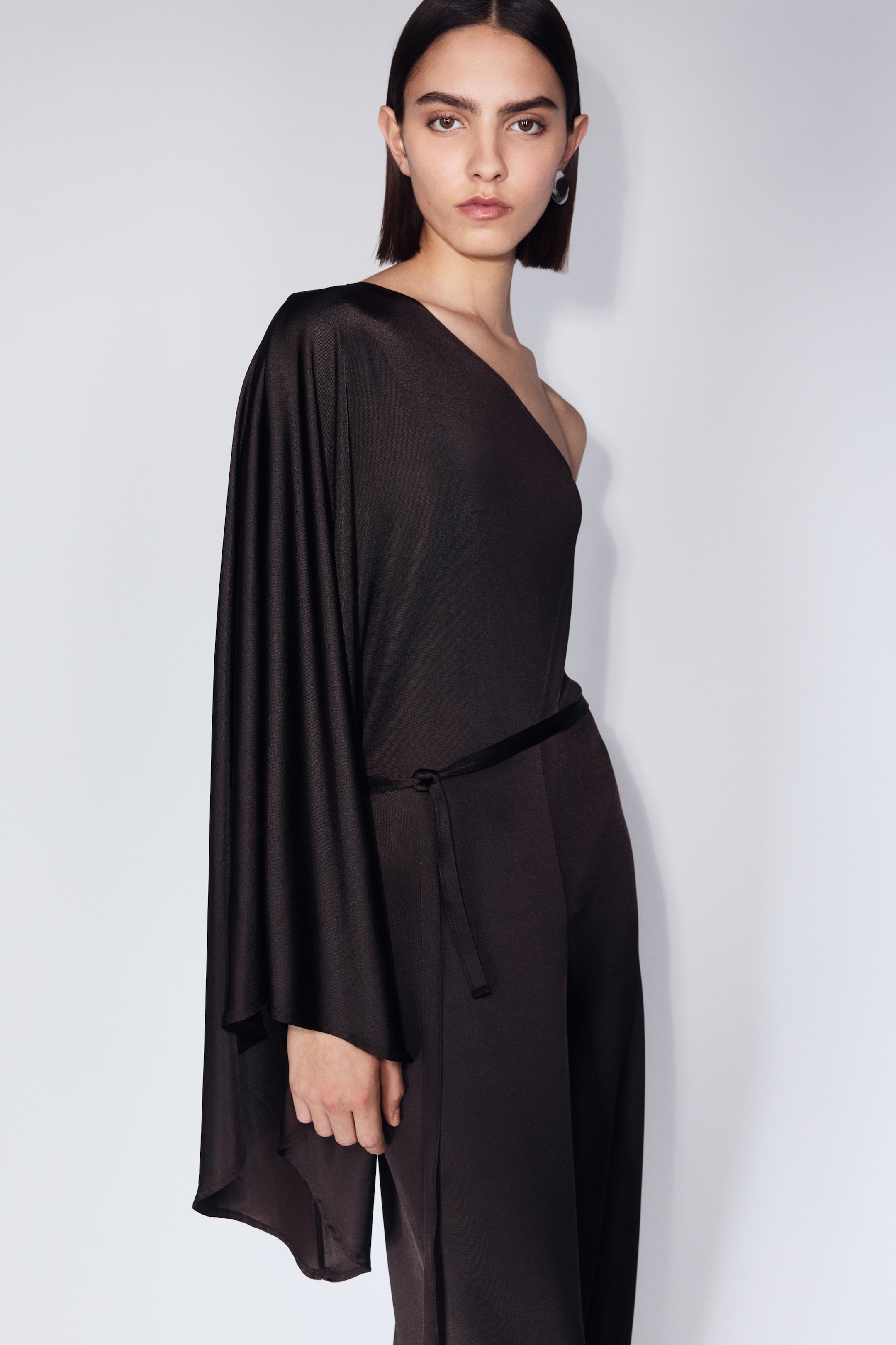 Vis større bilde: En mørk, asymmetrisk jumpsuit med én skulder har en lang, flytende erme på venstre side og en bar høyre skulder. Et tynt, mørkt knytebelte strammer inn midjen, mens det myke stoffet faller løst nedover kroppen.
