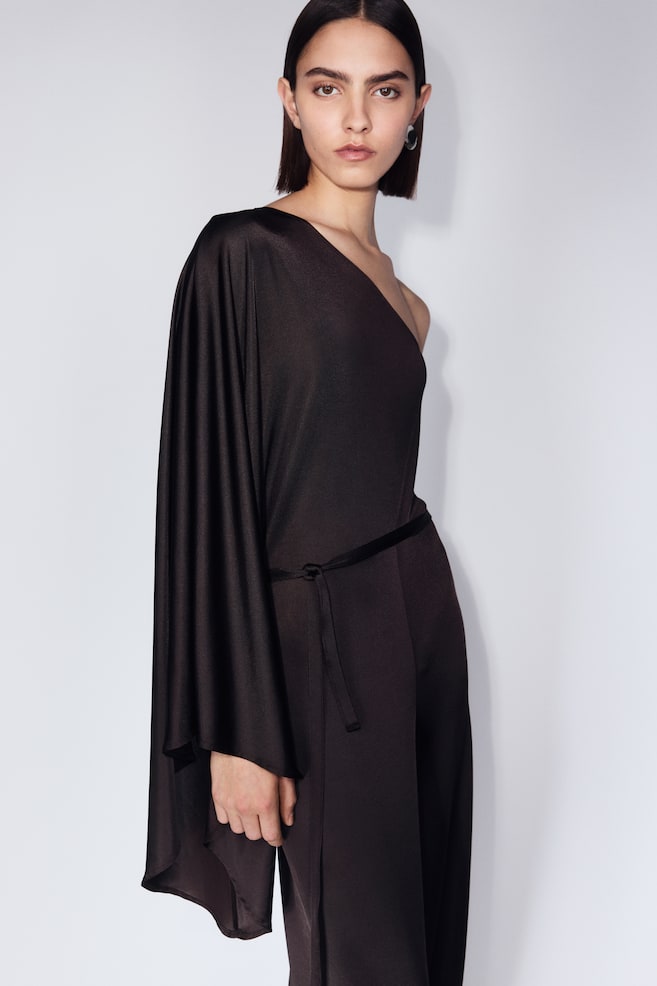 Ein dunkler One-Shoulder-Jumpsuit mit einem langen, fließenden Ärmel auf der linken Seite und einer freien rechten Schulter. Ein dünner, dunkler Bindegürtel betont die Taille, während der weiche Stoff locker am Körper herabfällt. - 1