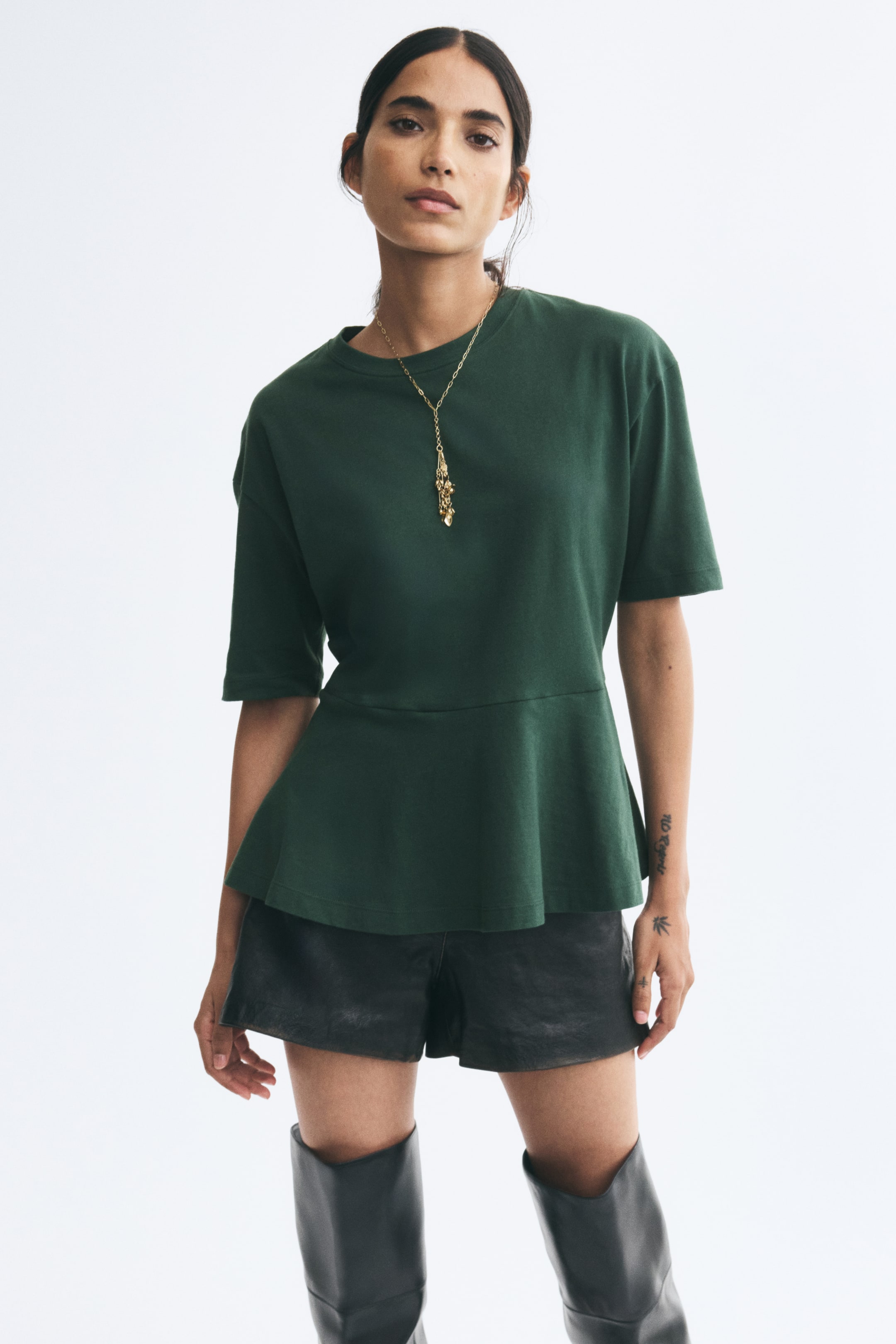 View larger image: A dark green cotton jersey top featuring a round rib-trimmed neckline, dropped shoulders, short sleeves, and a flared peplum is being worn by the model. The top is styled with black leather shorts, a gold pendant necklace, and tall black boots.
