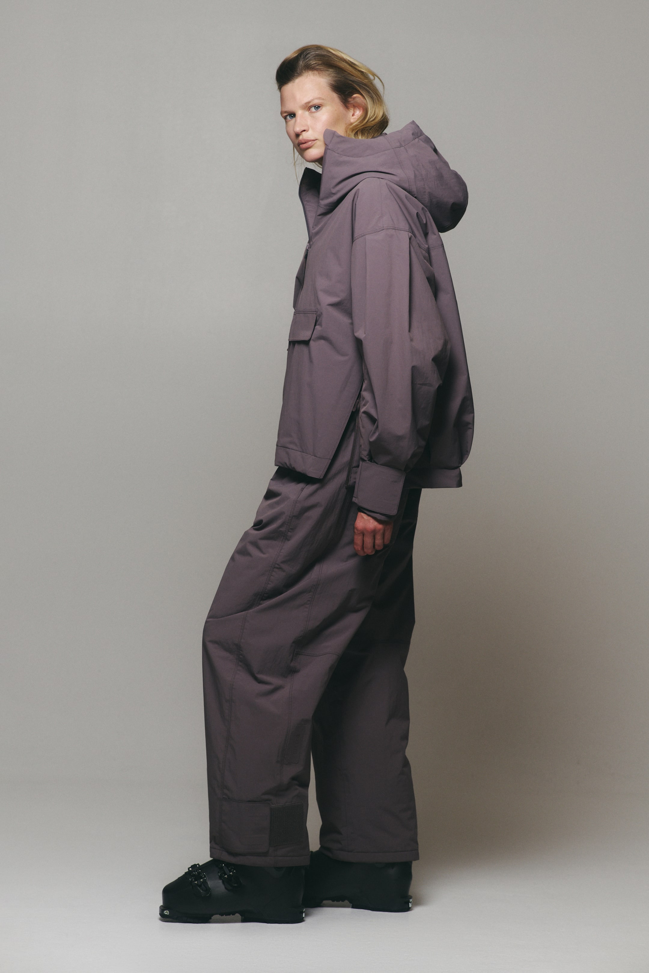 View larger image: A muted purple technical outerwear set, comprising a hooded pullover jacket with a front flap pocket and matching wide-leg pants, is worn by the model. The model stands in profile, paired with black ski boots.