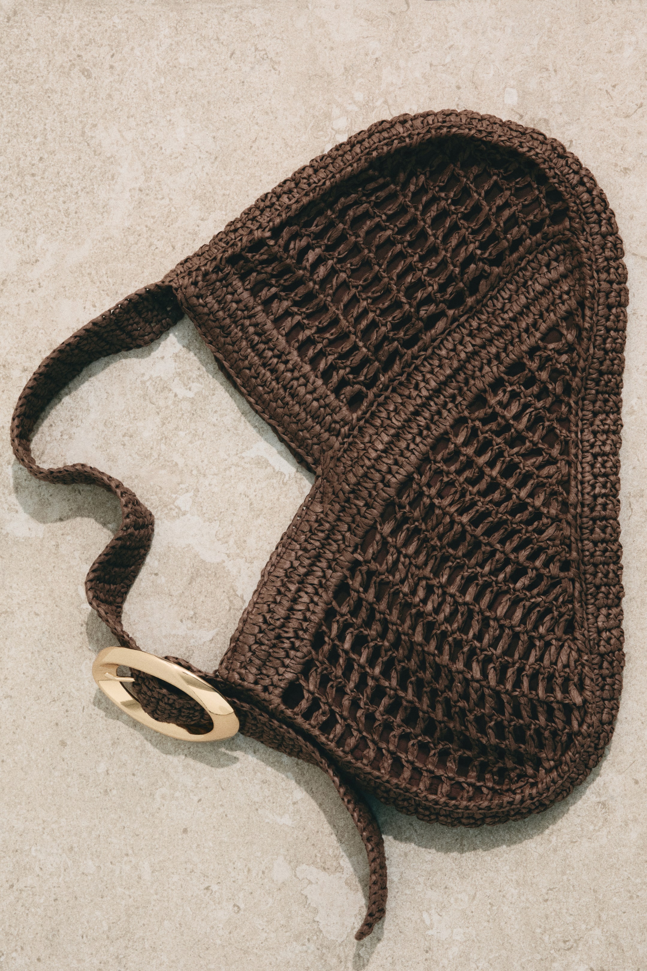 Agrandir l'image: Un sac en raphia tressé de couleur brun foncé est posé à plat, présentant un tissage ouvert en forme de filet qui laisse entrevoir une doublure en tissu plus sombre. Une boucle ovale dorée relie la sangle tressée au corps principal du sac.