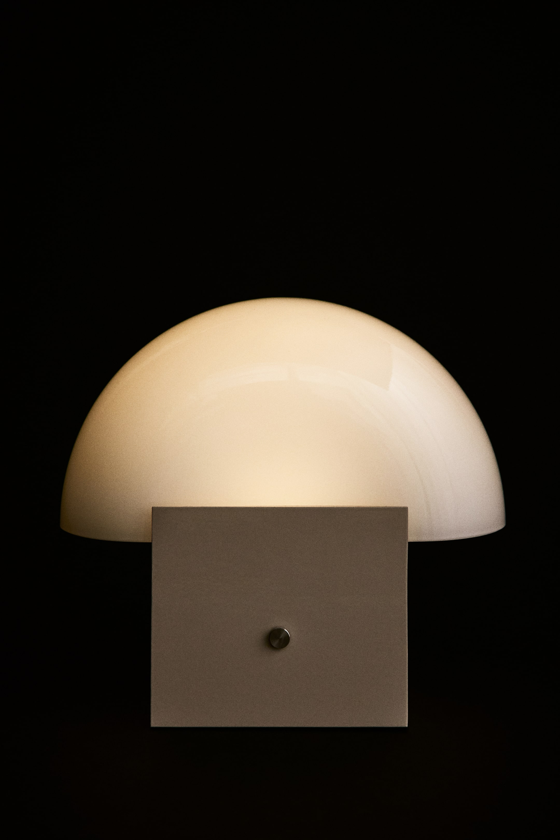Grösseres Bild ansehen: Eine wiederaufladbare Tischlampe mit geometrischem Design von vorne, die einen leuchtenden, gewölbten Glasschirm auf einem quadratischen Metallfuss zeigt. In der Mitte des Sockels befindet sich ein kleiner, runder Metallknopf.