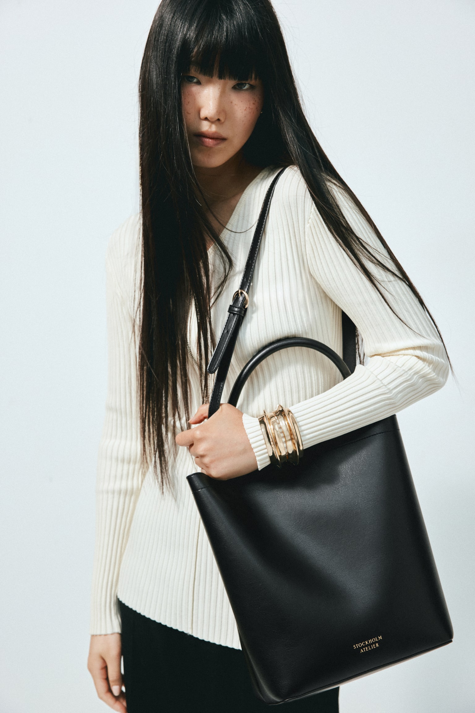 A black bucket bag is facing front, showcasing a smooth texture, a single curved top handle, and the 'STOCKHOLM ATELIER' logo in gold on its lower front. It features a concealed magnetic fastening at the top, a detachable and adjustable shoulder strap, and an internal divider creating two compartments, one of which is larger than the other, creating a slouchy effect; it is also lined. - 1
