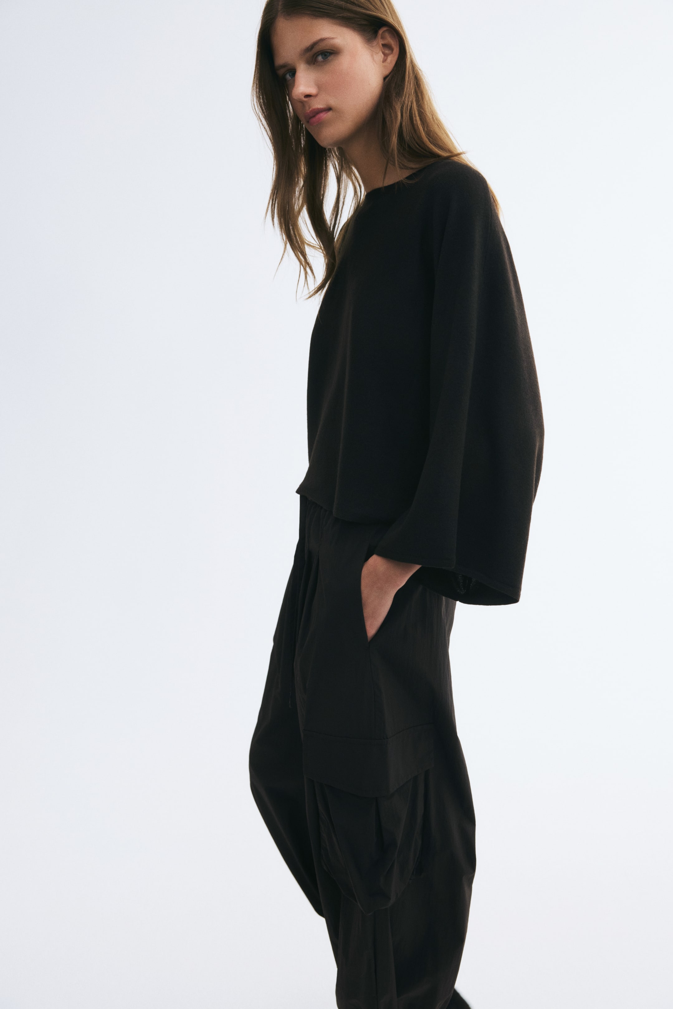 View larger image: The model is wearing a short, loose-fit black knit top with a boat neck and wide, long cape sleeves. The top is styled with loose-fitting black trousers, with one hand resting in a pocket.