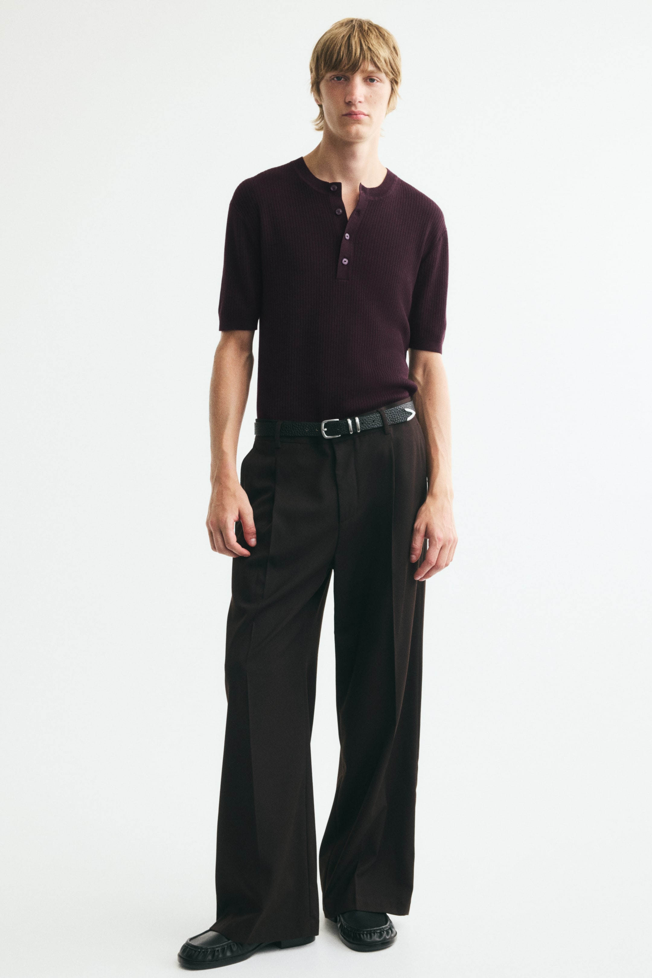 View larger image: A dark purple ribbed short-sleeve henley shirt with a buttoned placket is worn by the model. The model pairs it with dark brown wide-leg trousers and a black belt.