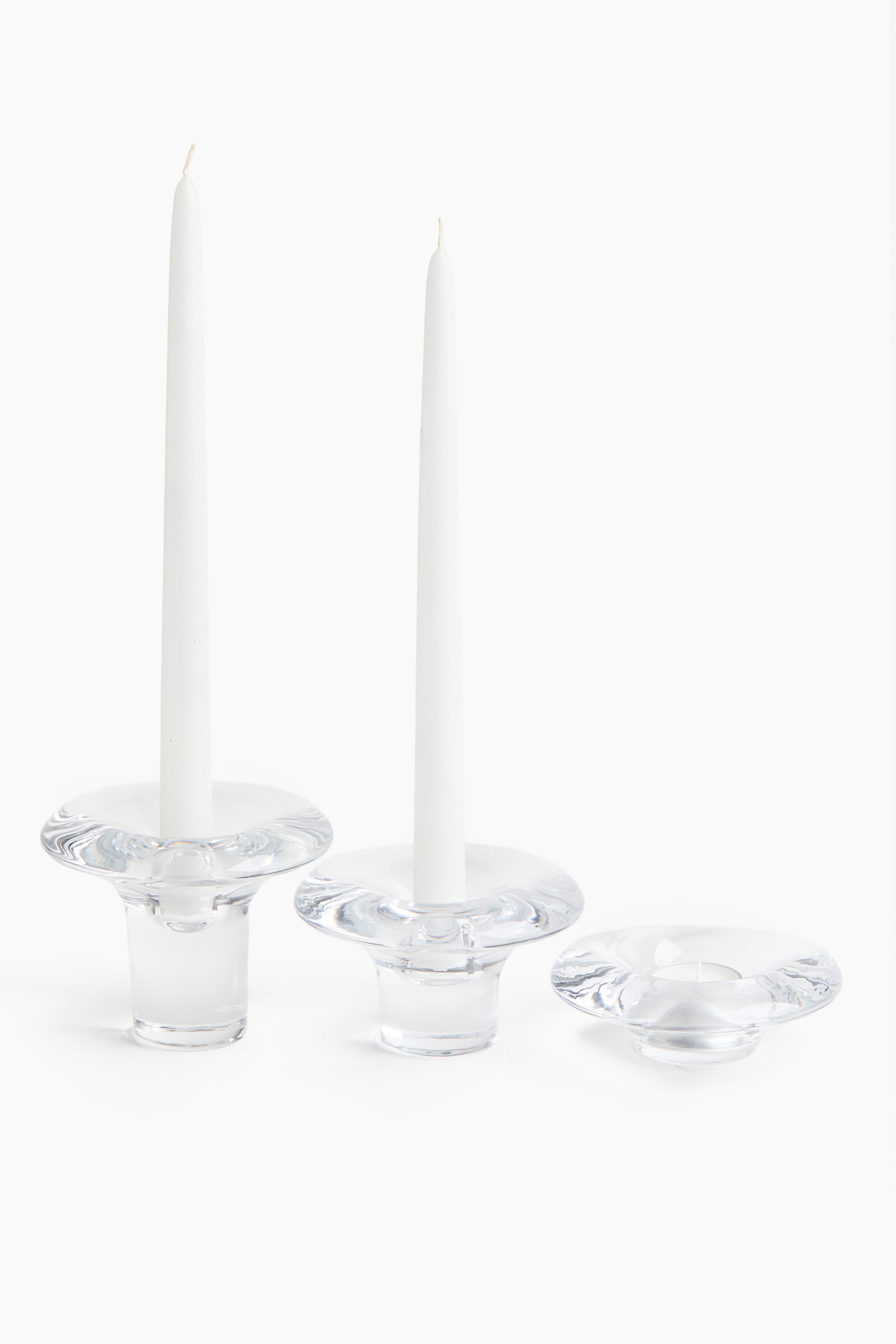 View larger image: Three clear glass candle holders are standing in varying heights, each featuring a flared top and smooth, curved surfaces. The two taller holders are presenting white taper candles, while the shortest holder is containing a white tealight.