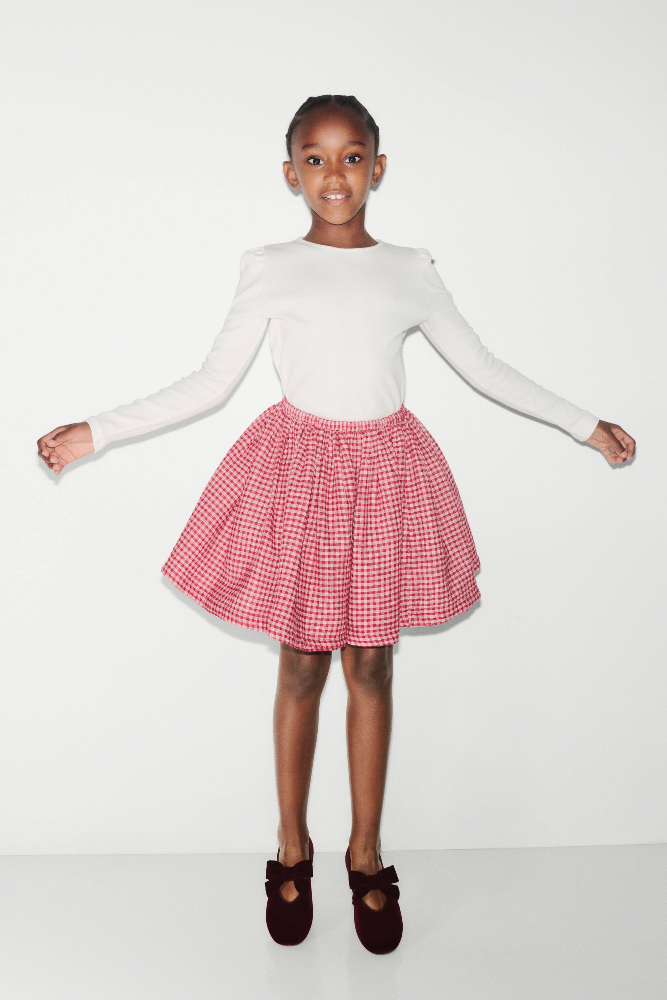 View larger image: A red and white gingham skirt is shown from the front, featuring a gathered, elasticised waistband that creates a flared, knee-length silhouette. The skirt is styled with a tucked-in white long-sleeve top.