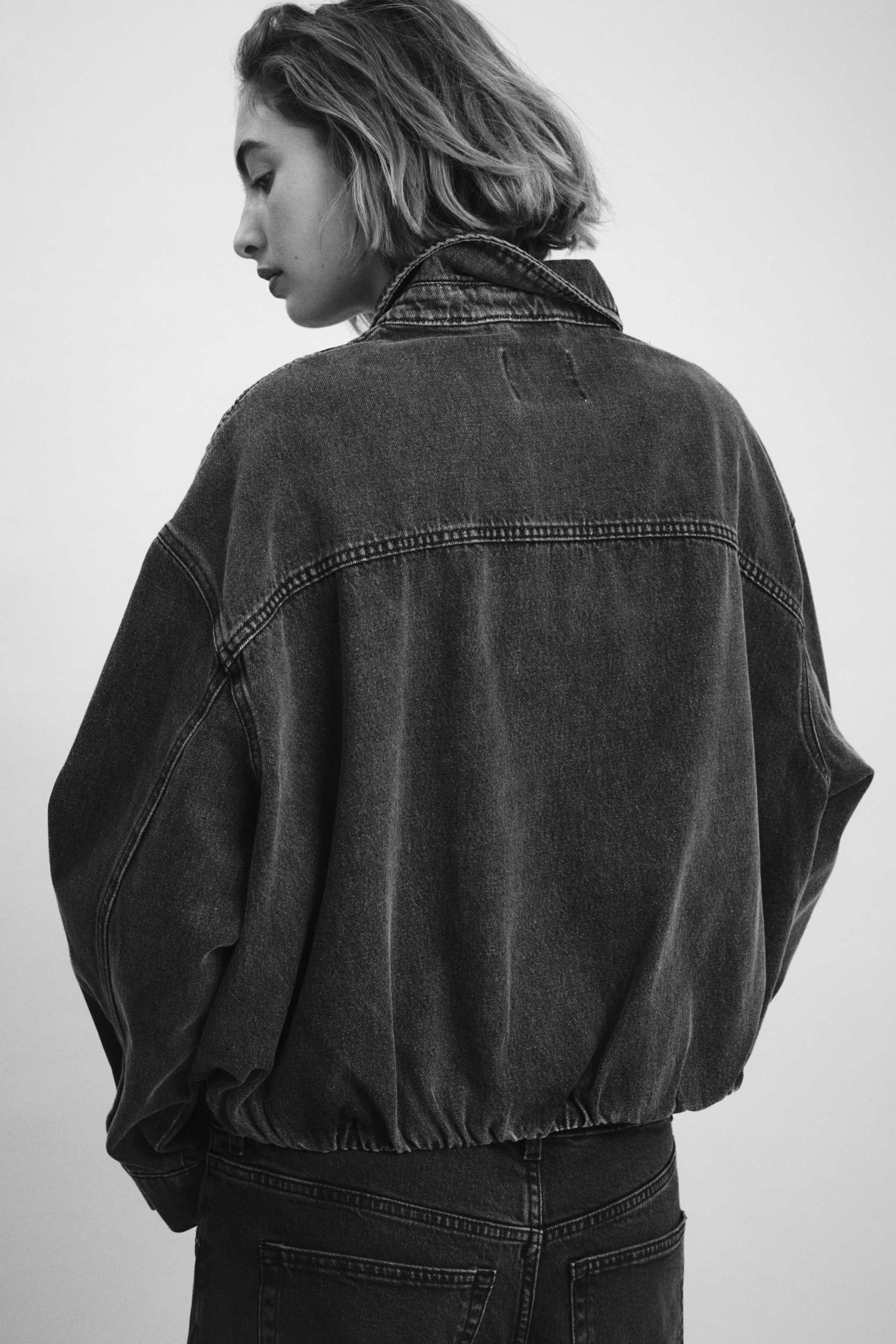 Προβολή μεγαλύτερης εικόνας: Ένα τζάκετ από τζιν με σκούρο ξέπλυμα φαίνεται από την πίσω πλευρά, διαθέτοντας μια υπερμεγέθη εφαρμογή με χαμηλωμένους ώμους, κλασικό γιακά, οριζόντια ραφή στην πλάτη και ελαστική μέση με σούρες.