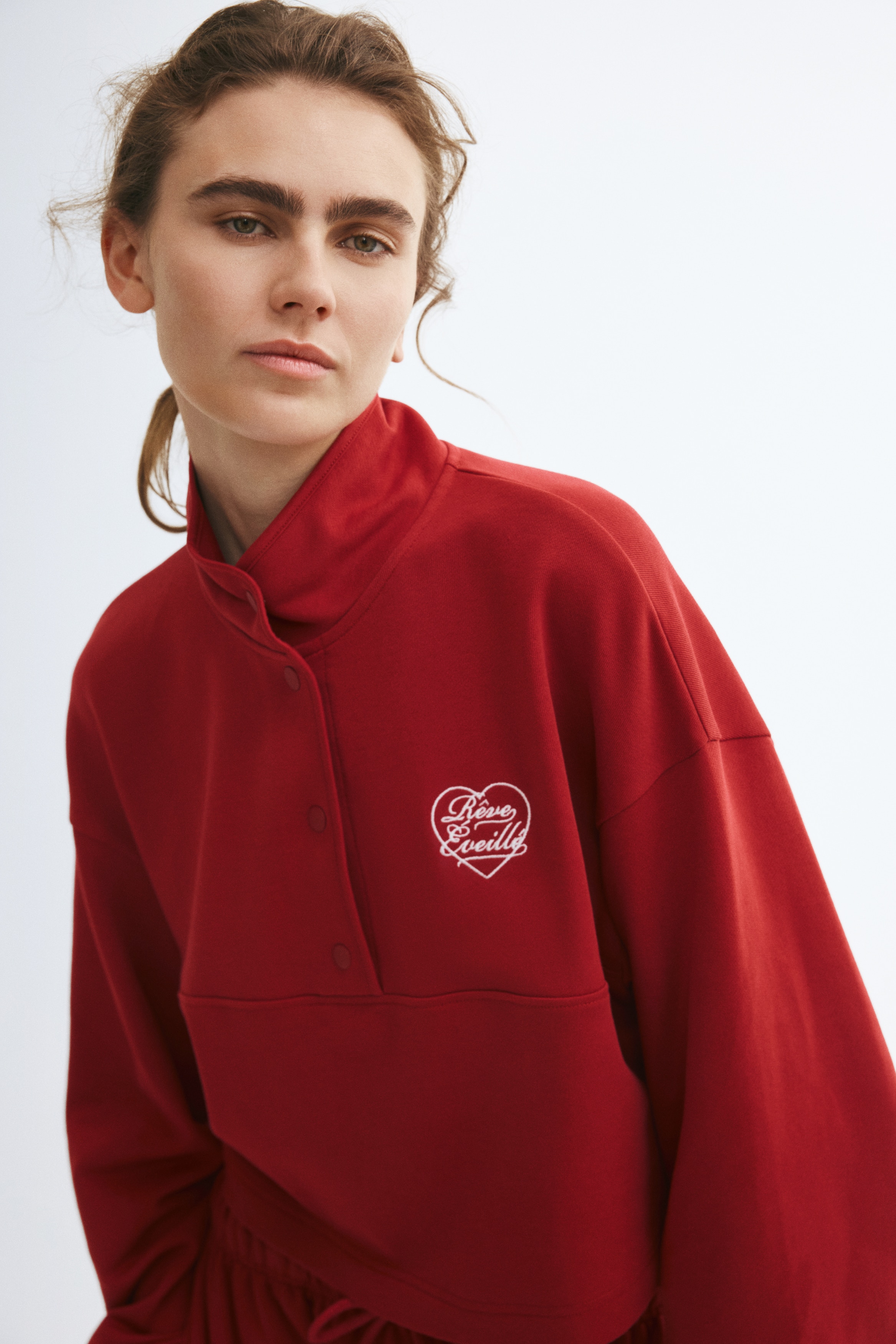 The red cropped sweatshirt is shown from the front, featuring a mock neck with a snap-button placket and dropped shoulders. It displays a small white embroidered heart graphic on the left chest and is finished with elasticated cuffs on the long sleeves.