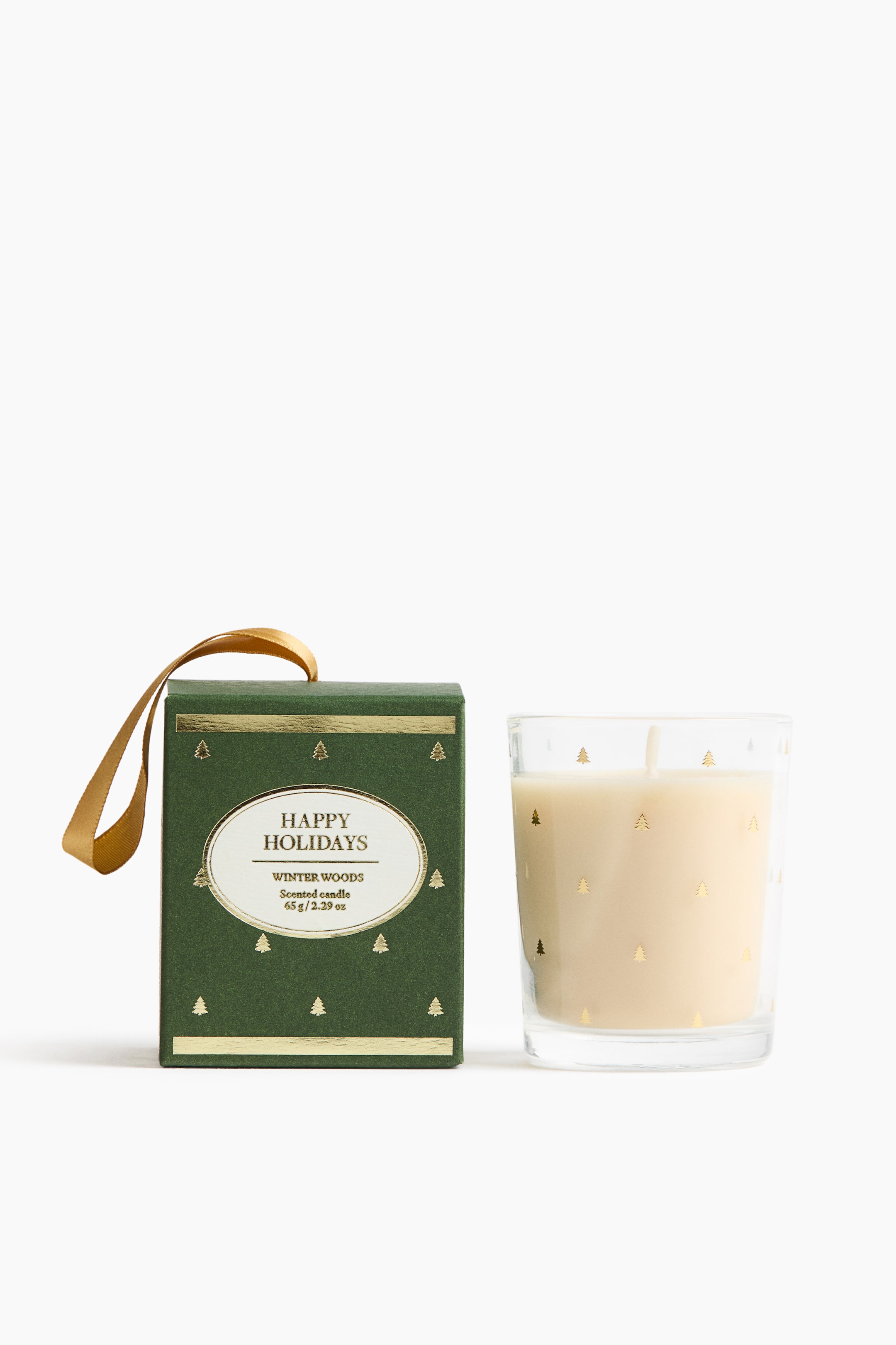 Ampliar la imagen: Una vela aromática en un vaso de vidrio transparente, detallado con pinos en lámina dorada, está junto a su caja verde oscuro a juego. La caja, orientada al frente, presenta un lazo de cinta dorada en la parte superior y una etiqueta ovalada blanca al centro que dice "HAPPY HOLIDAYS" y "WINTER WOODS".