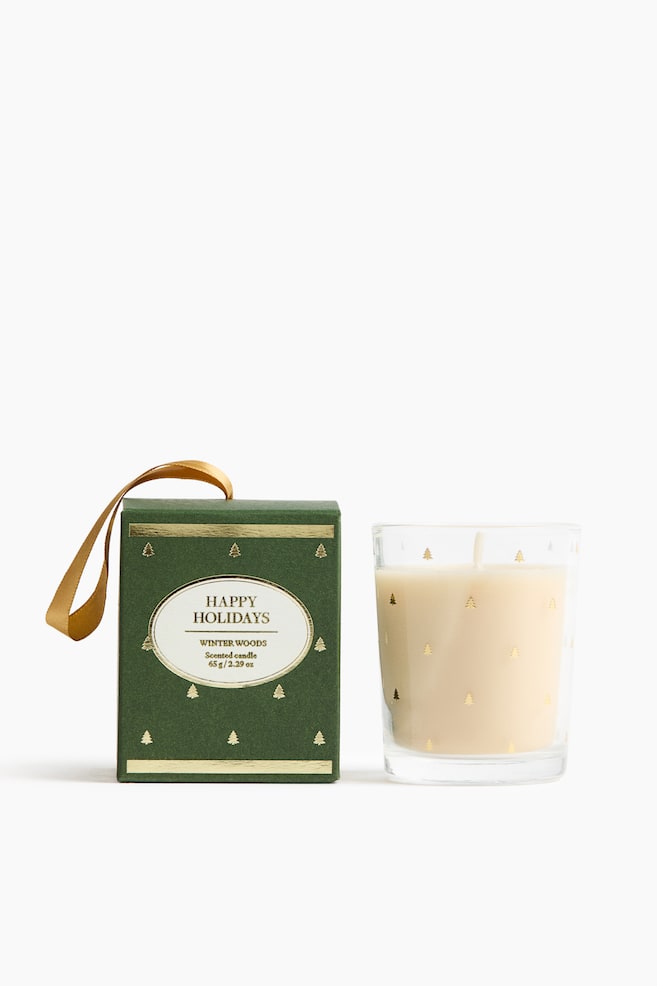A scented candle in a clear glass tumbler, detailed with gold-foil pine trees, stands next to its matching dark green box. The front-facing box features a gold ribbon loop on top and a central white oval label reading "HAPPY HOLIDAYS" and "WINTER WOODS". - 1