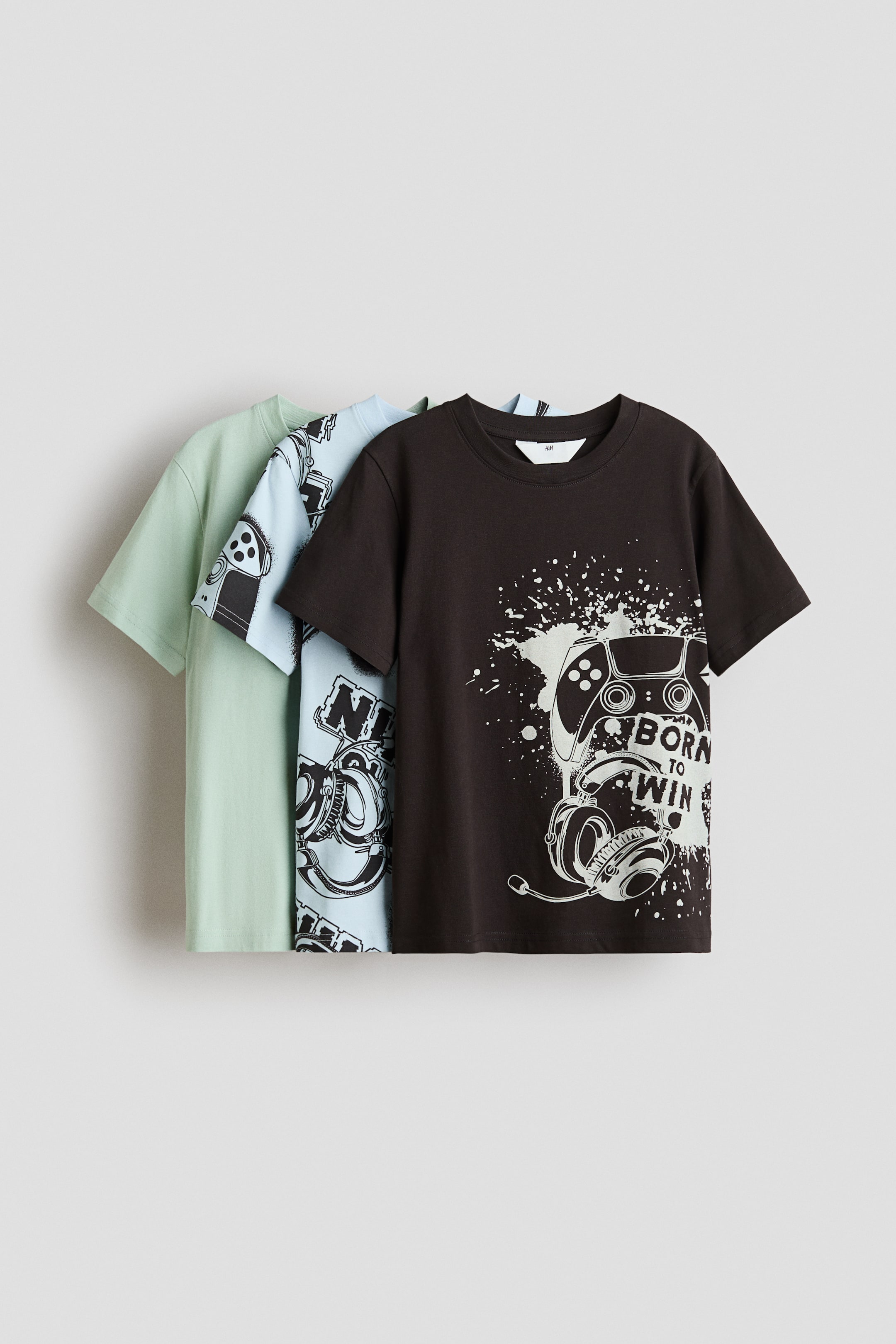 View larger image: Three t-shirts are showcased, layered front-facing. The foremost dark brown t-shirt displays a white gaming controller and headphones graphic, while the middle light blue t-shirt features an all-over gaming controller pattern. The rearmost light green t-shirt appears solid. All t-shirts are designed with a round, rib-trimmed neckline and gently dropped shoulders.