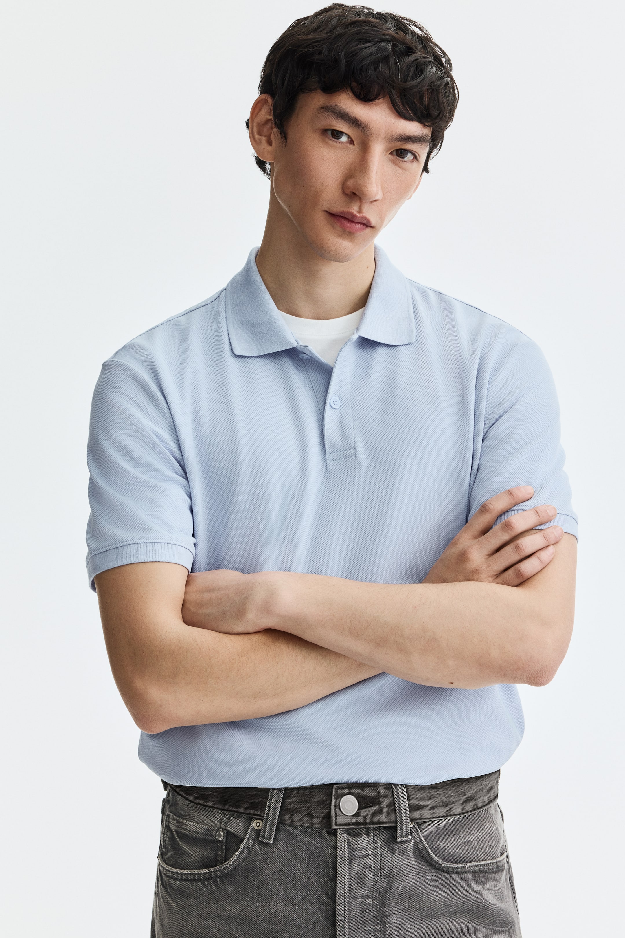 Voir une image plus grande: Un polo bleu clair présente une texture en maille piquée, un col pointu et une patte de boutonnage à deux boutons de couleur claire. Il est conçu avec des manches courtes et des poignets côtelés, et est porté face avant sur un t-shirt à col rond blanc.