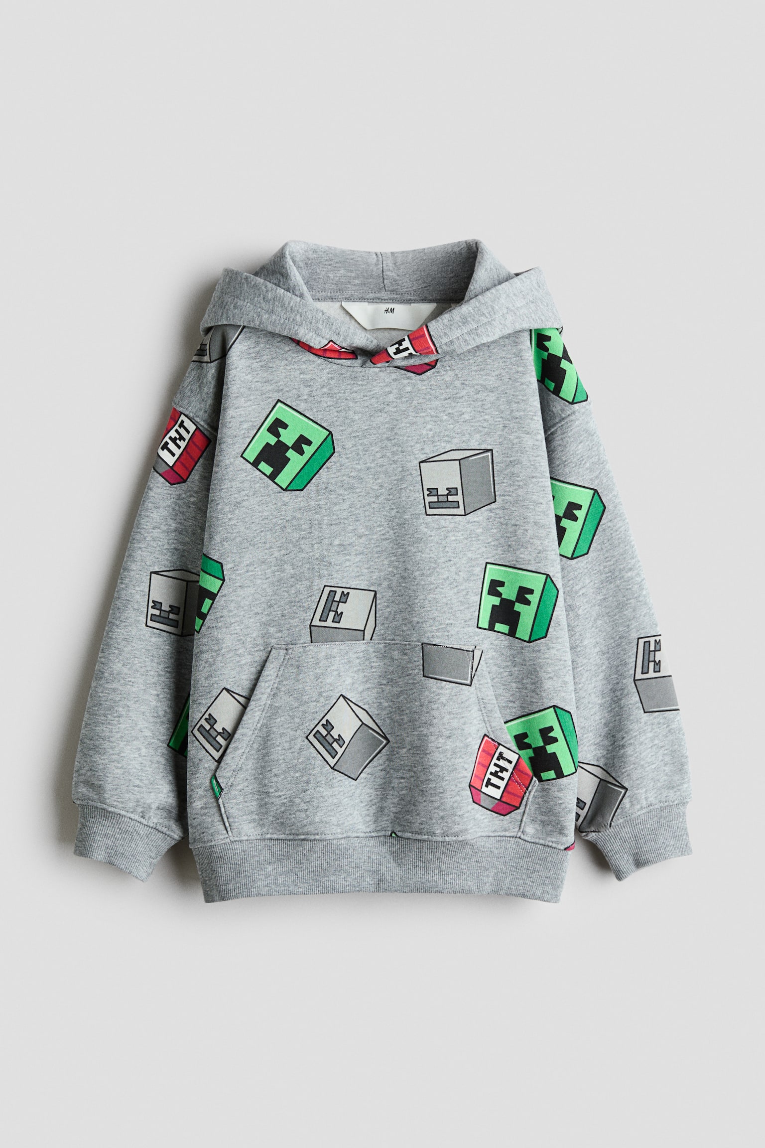 A loose-fit, light gray fleece hoodie is facing front, showcasing an all-over print of pixelated block graphics including green Creepers, Skeletons, and TNT. The garment is displaying a wrap-front hood, dropped shoulders, a front kangaroo pocket, and ribbed cuffs and hem.