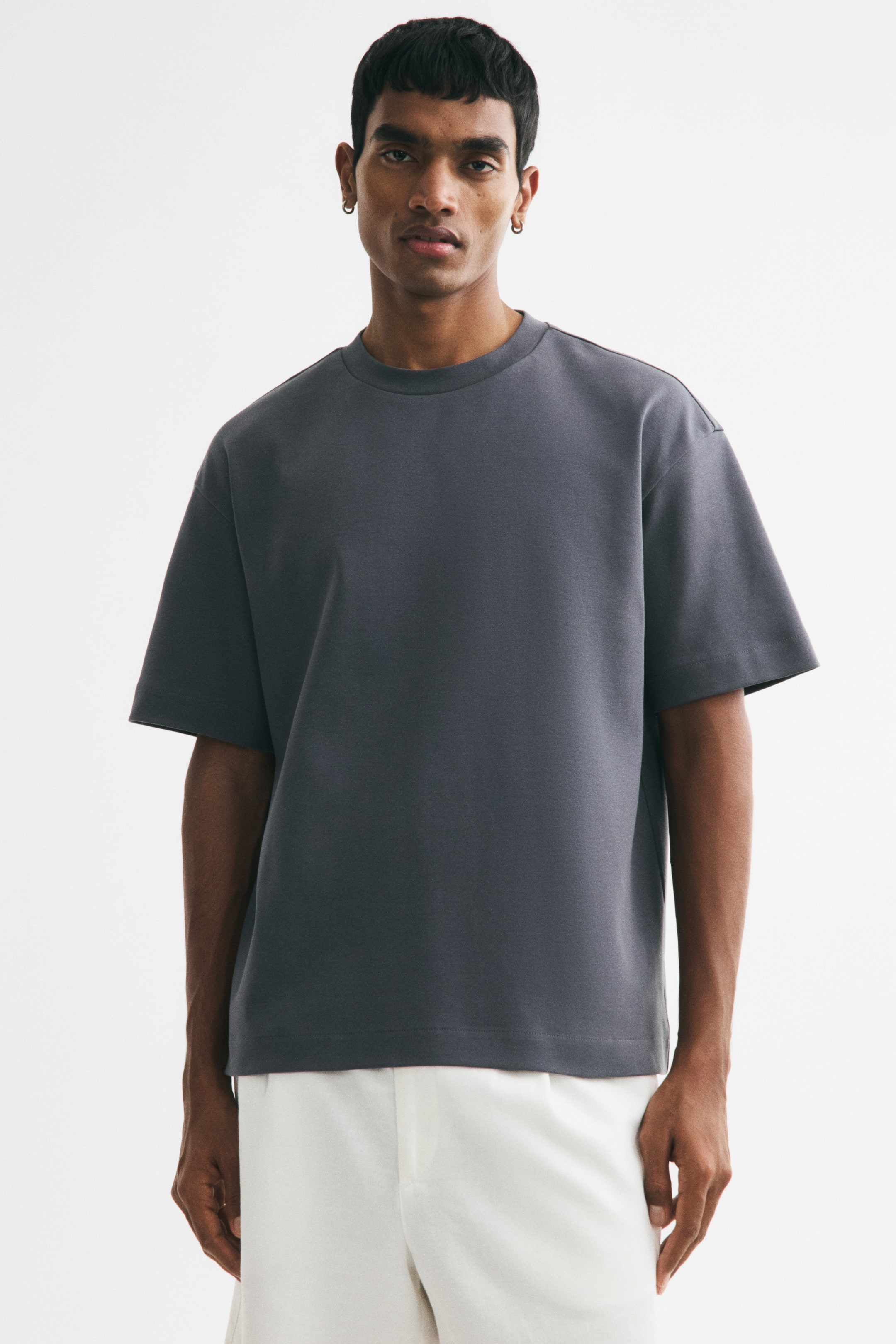View larger image: The model is wearing a dark gray, boxy-fit t-shirt, showcasing its ribbed crew neckline, dropped shoulders, and straight hem. The loose-fitting tee is styled with white trousers.