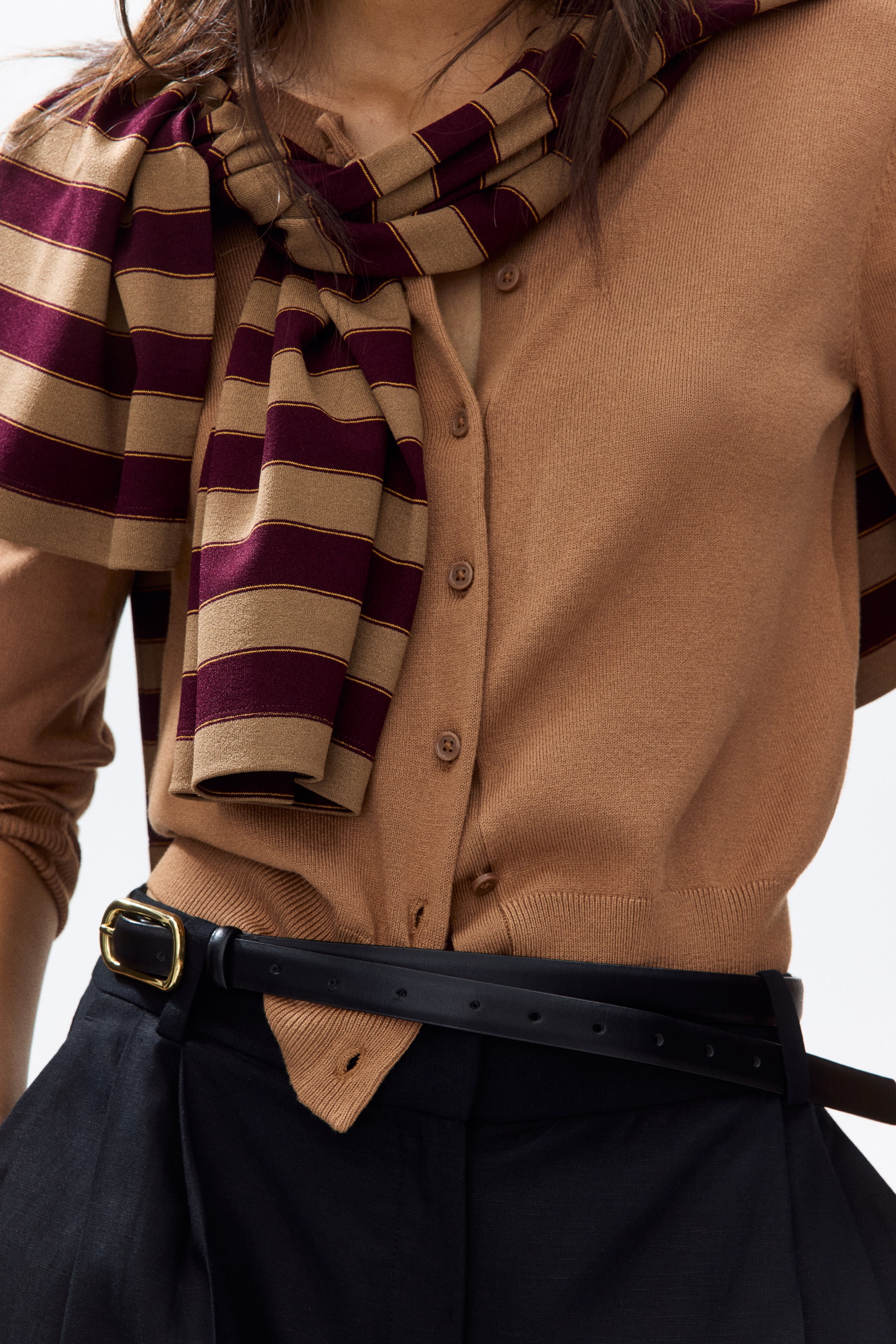 View larger image: The model is wearing a camel-colored fine-knit viscose blend cardigan, which features a round, rib-trimmed neckline, front buttons, and ribbing at the hem. Draped over the cardigan is a striped scarf, paired with black trousers and a belt underneath.