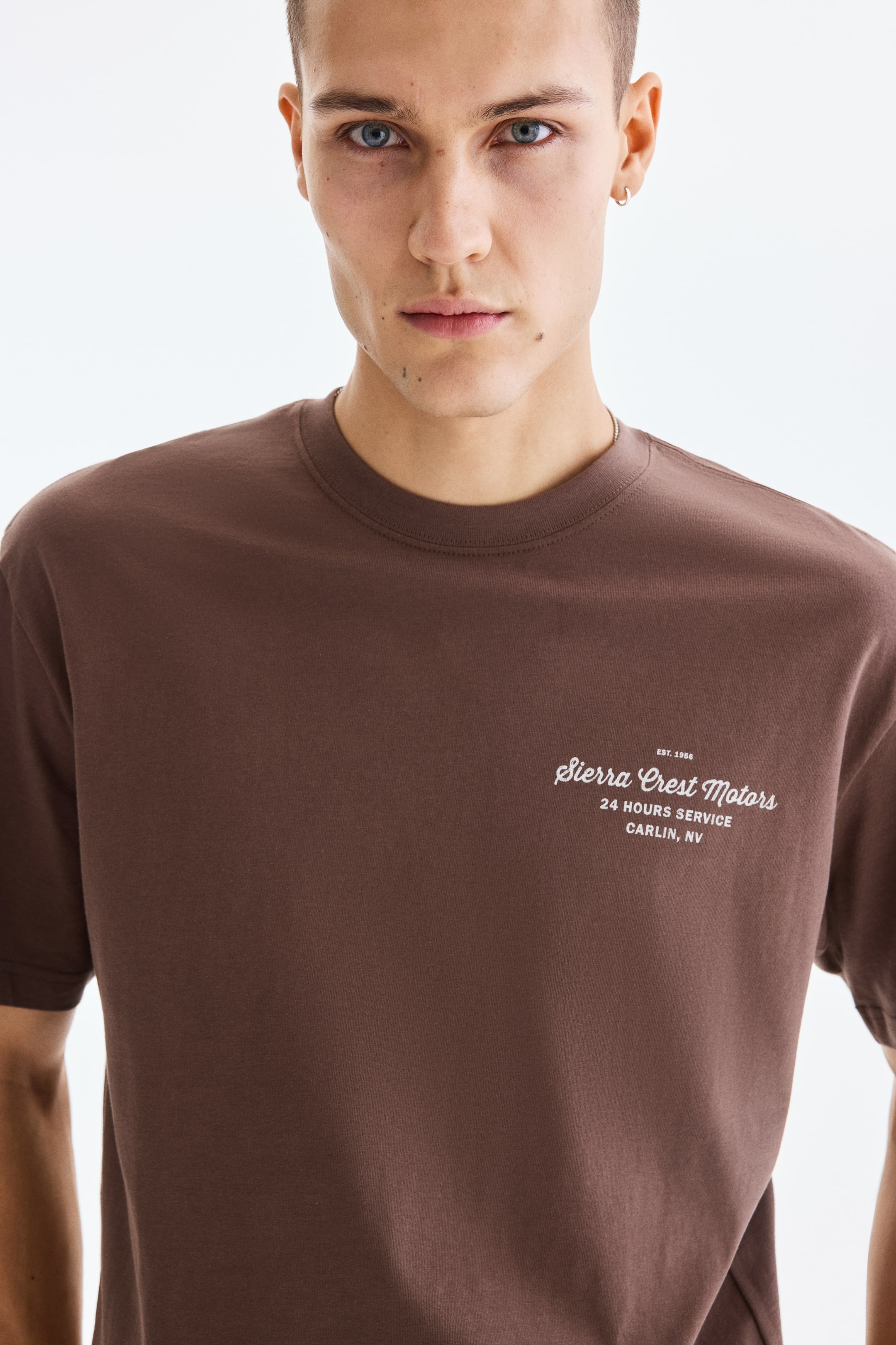View larger image: A brown crew neck t-shirt showcases a white graphic print on its front right chest. The print displays "EST. 1956 Sierra Crest Motors 24 HOURS SERVICE CARLIN, NV".