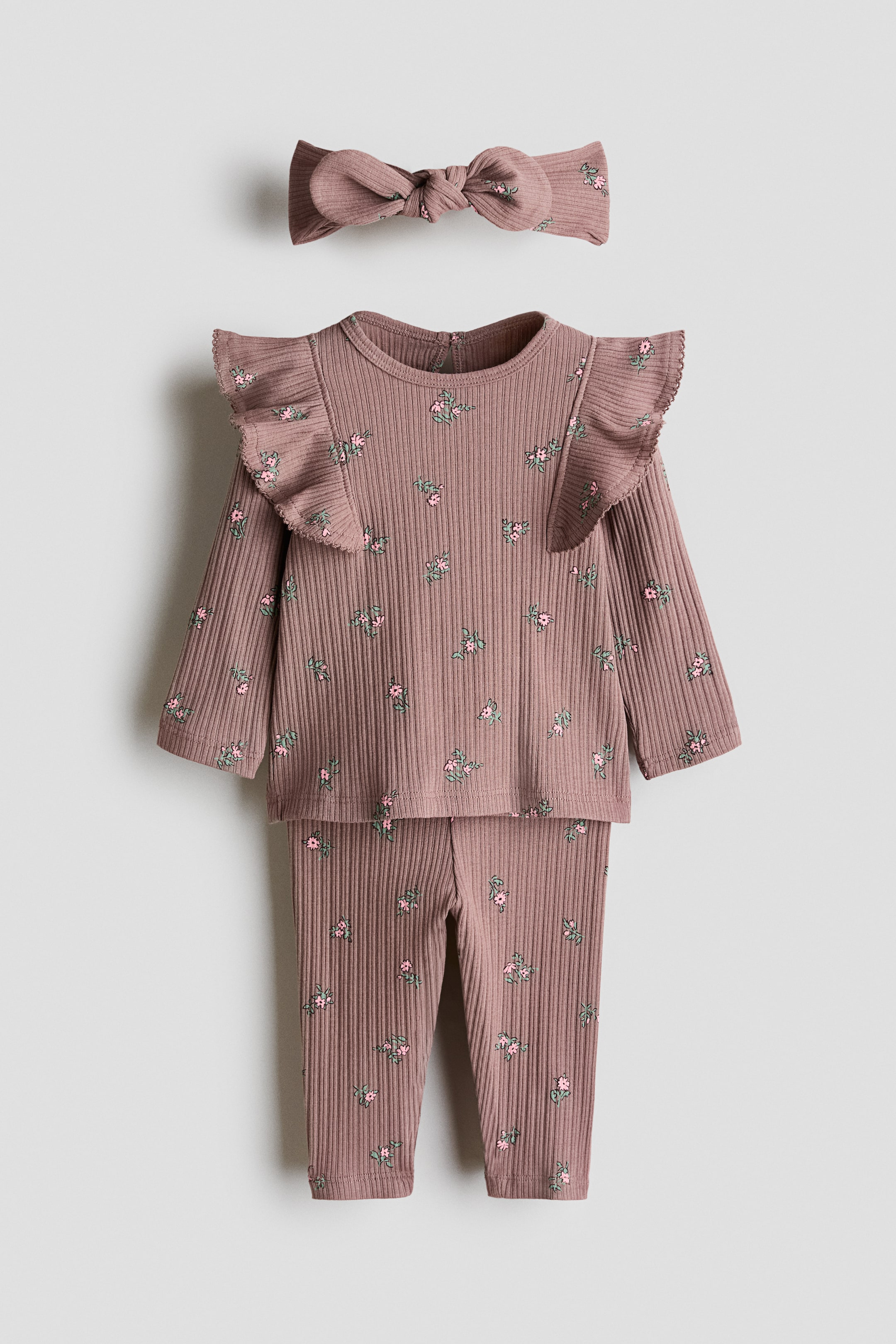 View larger image: A baby outfit set is showcased front-facing, comprising a long-sleeved top, long pants, and a coordinating bow headband. The ribbed, dusty rose fabric, embellished with a subtle pink and green floral print, features ruffles on the top's shoulders and a snap fastening at the back of its round neck.