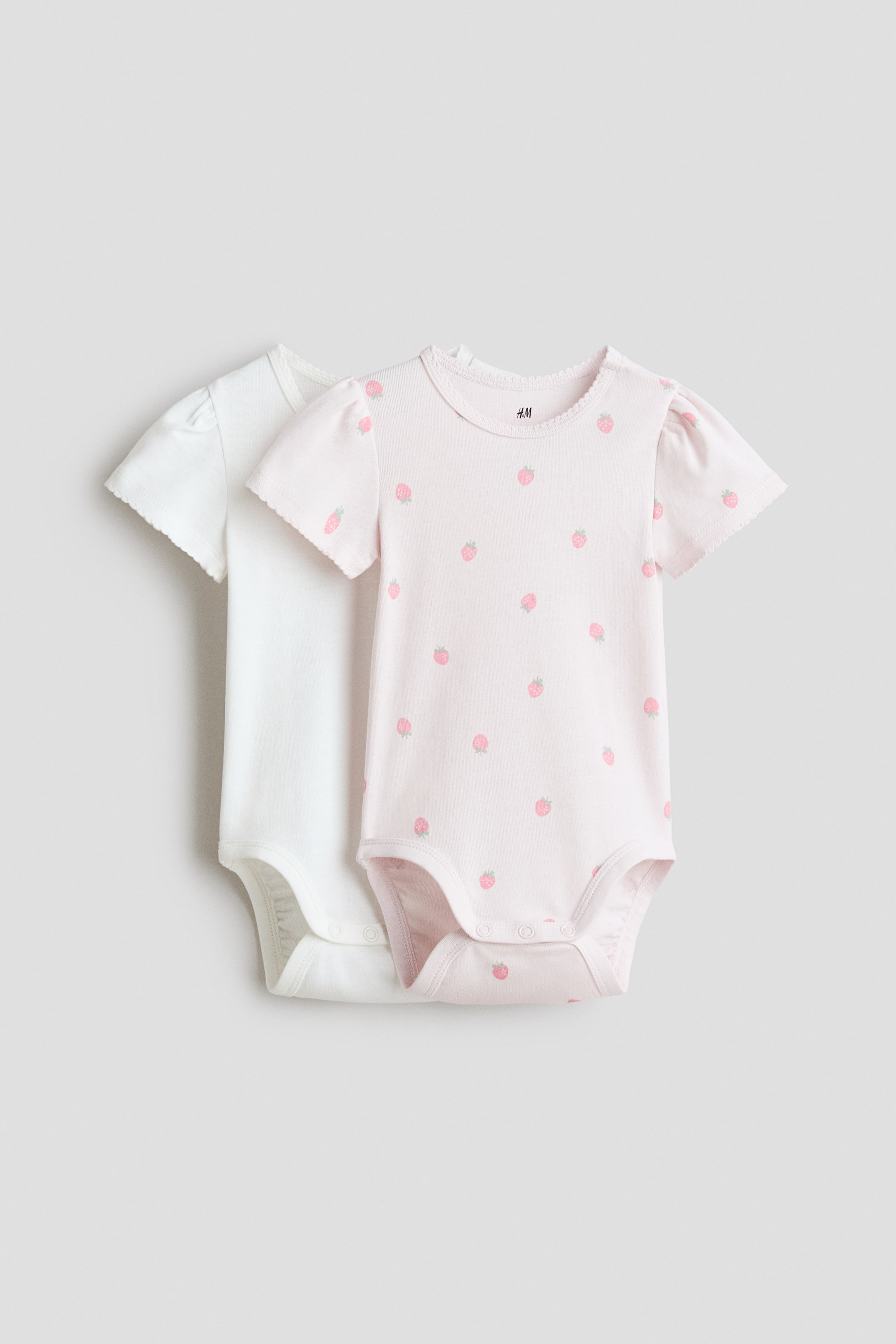 Visualizza immagine più grande: Due body a maniche corte sono presentati in PRIMO PIANO. Il body davanti è rosa chiaro con un motivo ripetuto di fragole, mentre quello dietro è bianco. Entrambi i body presentano maniche corte leggermente a sbuffo con bordo smerlato, uno scollo rotondo smerlato, bottoni automatici al cavallo e cuciture arricciate intorno alle gambe.