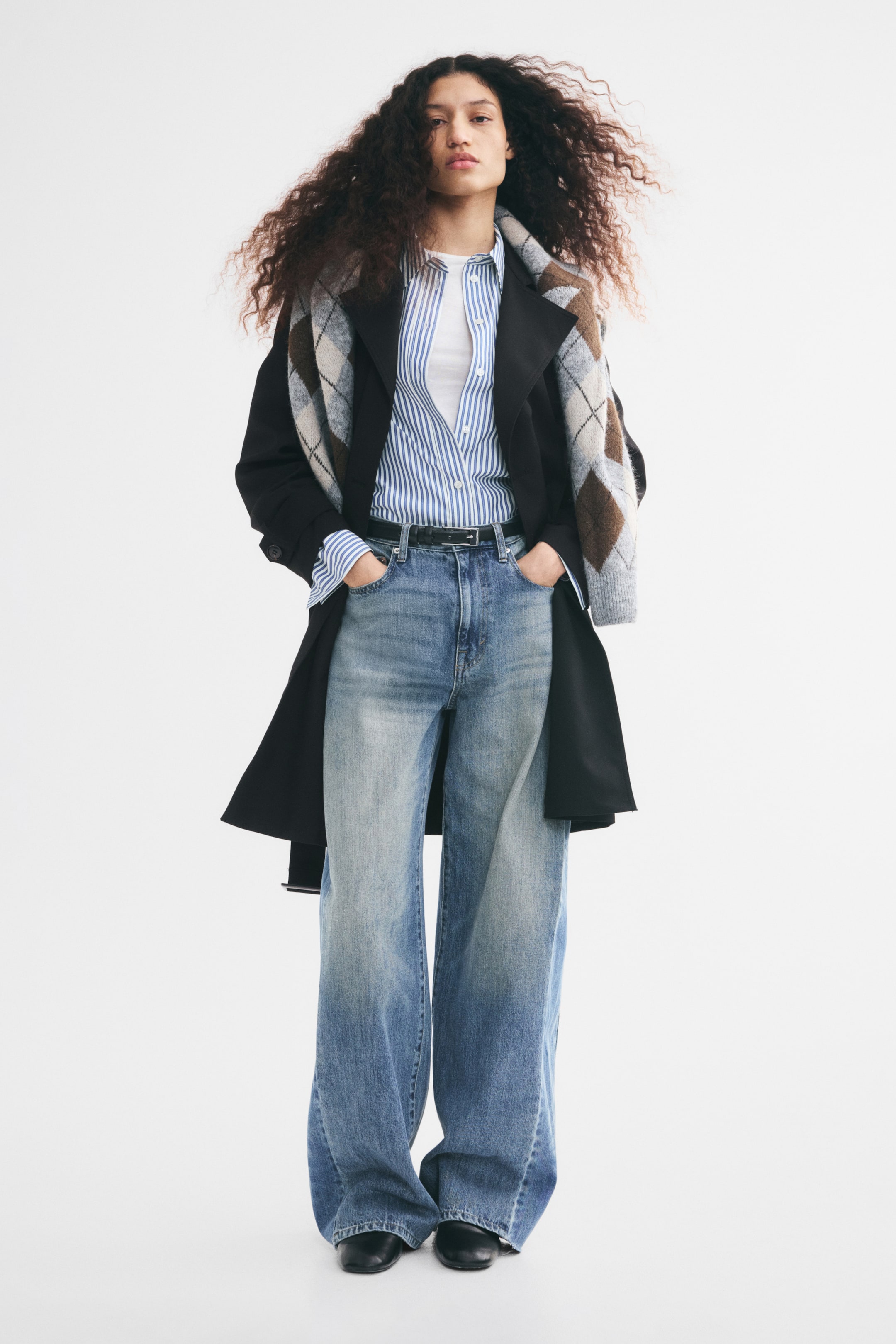 View larger image: A black, knee-length trench coat is worn open by the model, layered over a blue-and-white striped button-down shirt and wide-leg denim jeans. The coat features wide notch lapels and is styled with a grey and brown argyle knit sweater draped over the shoulders.