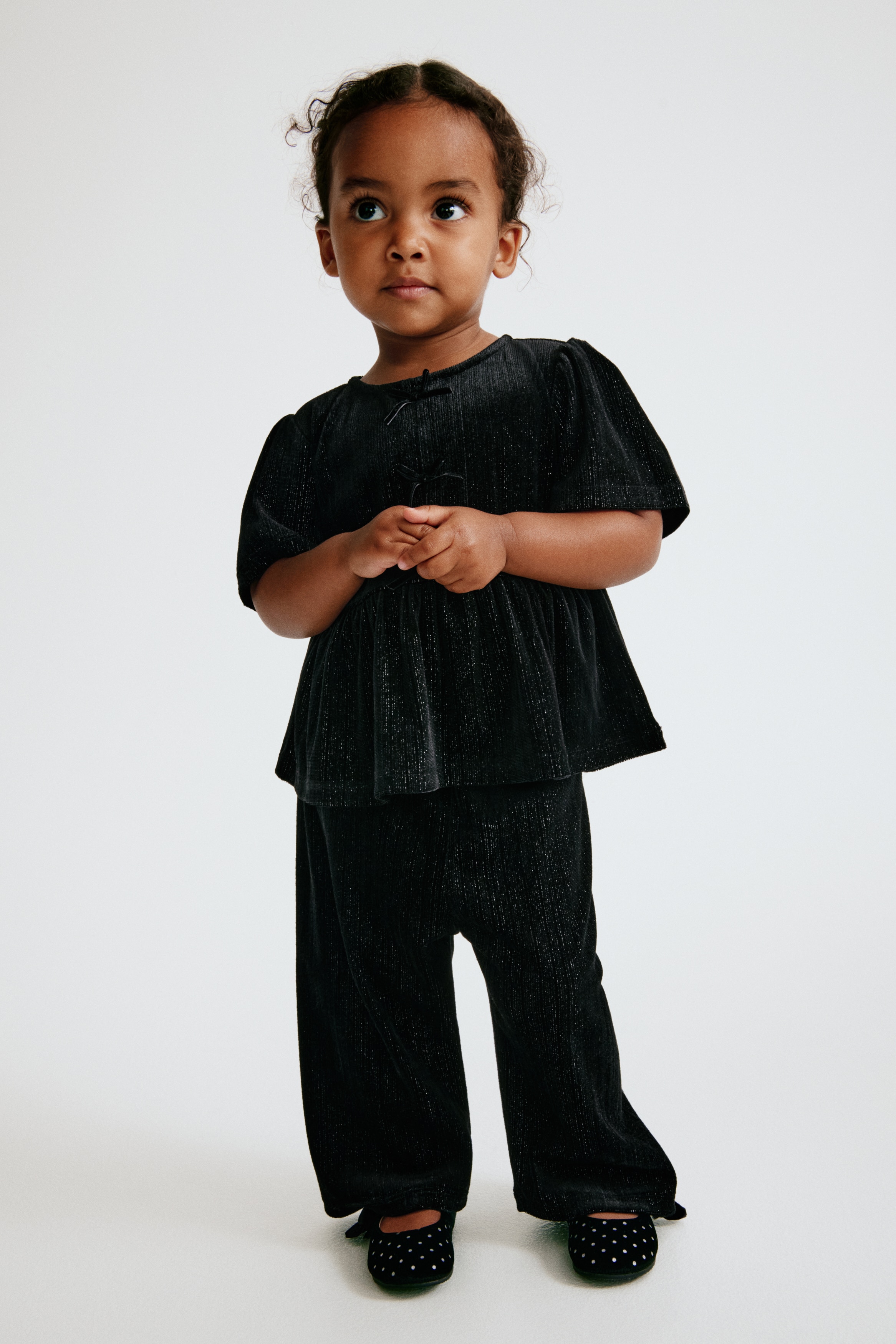 A two-piece black clothing set, shown from the front, featuring a short-sleeved peplum top and matching straight-leg trousers crafted from a textured, subtly shimmering velvet fabric. The top features a round neckline, slightly puffed sleeves, and three small decorative bows down the centre bodice.