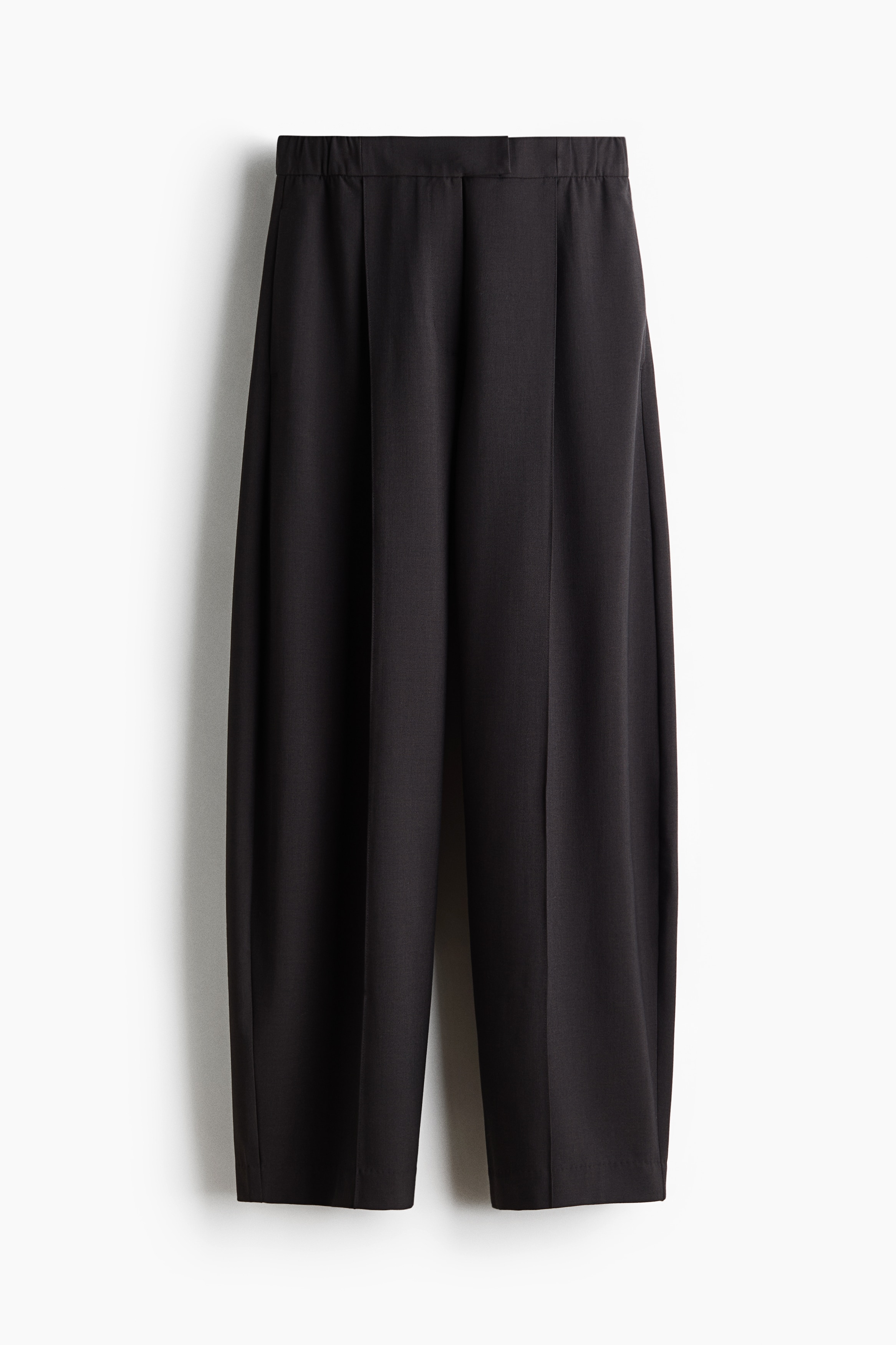 A model is wearing the loose-fit dark brown woven trousers, showcasing the front design. The trousers feature a regular waist with an extended waistband, front pleats, and sharp vertical creases extending down the wide, curved barrel legs.