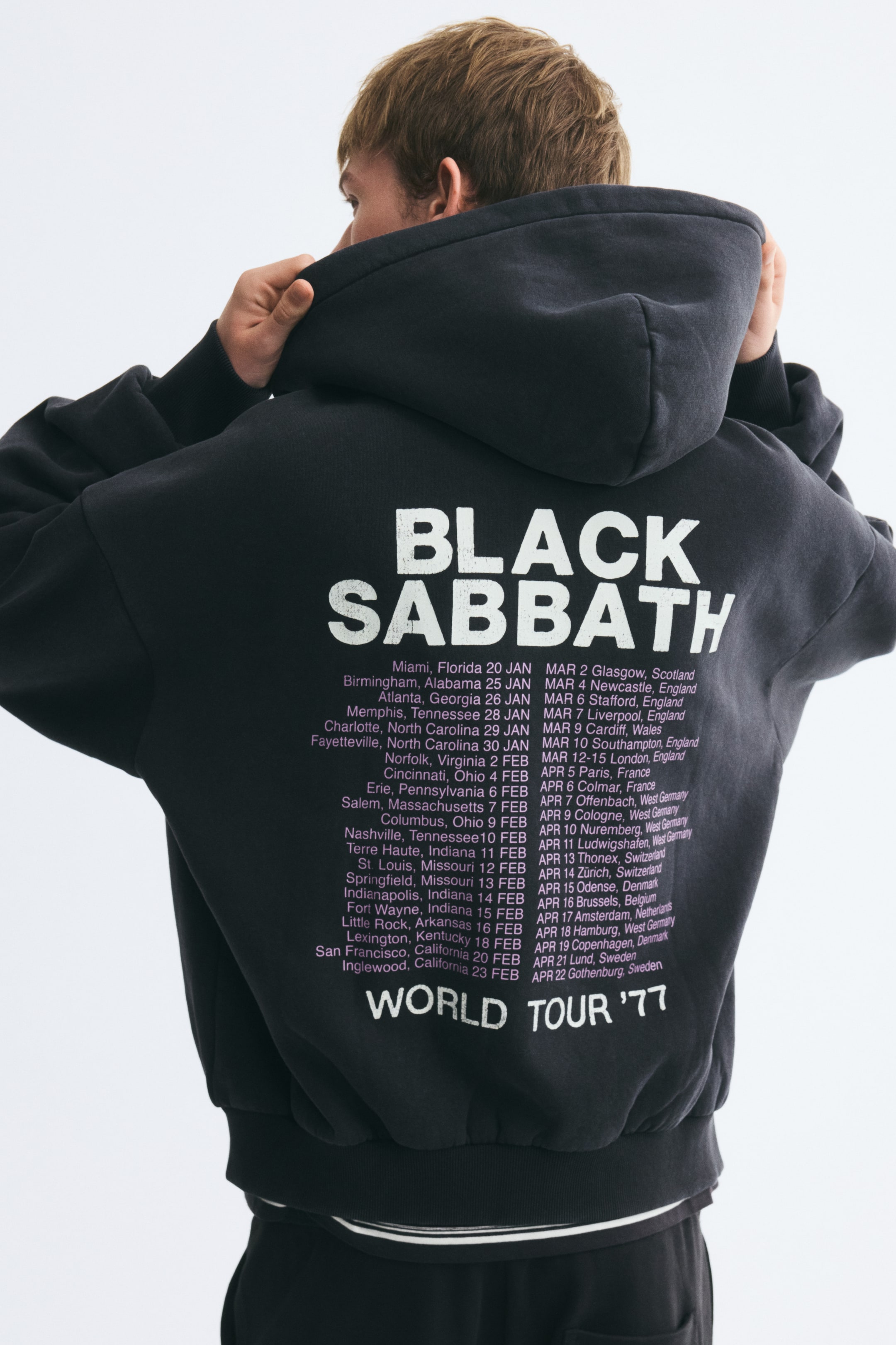View larger image: A black oversized hoodie featuring a white "BLACK SABBATH WORLD TOUR '77" graphic print on the back is being worn by the model, who is viewed from behind. The model is adjusting the hood with both hands, showcasing the relaxed fit, dropped shoulders, and ribbed cuffs of the garment.