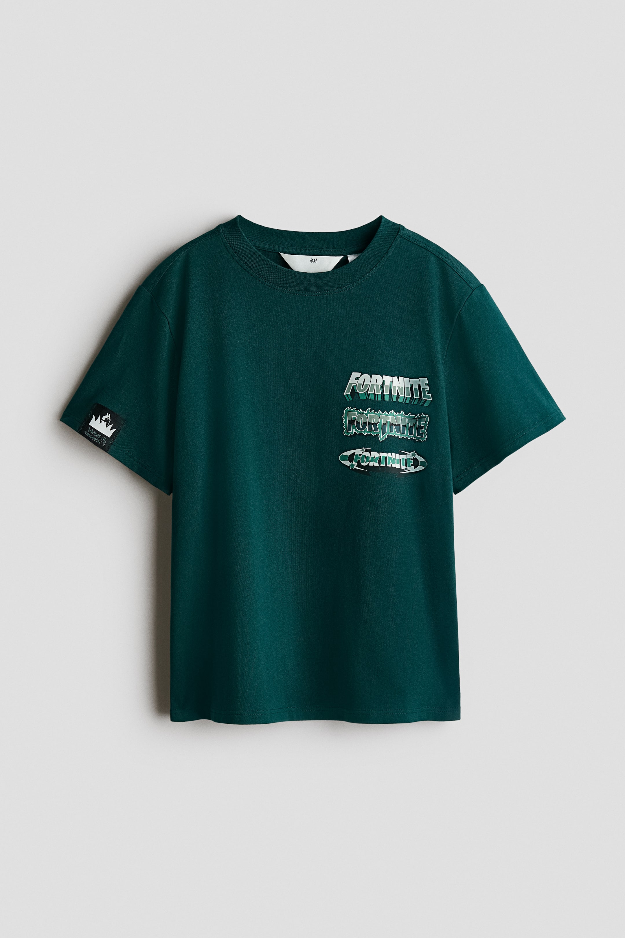 拡大画像を表示: ダークグリーンのコットンジャージーTシャツがその前面を見せており、丸いリブ付きネックラインとストレートカットの裾が特徴です。前面にはメタリックグリーンとシルバーのトーンで描かれた3つの重なった「Fortnite」プリントモチーフが飾られており、左袖には小さな黒いグラフィックパッチも見られます。