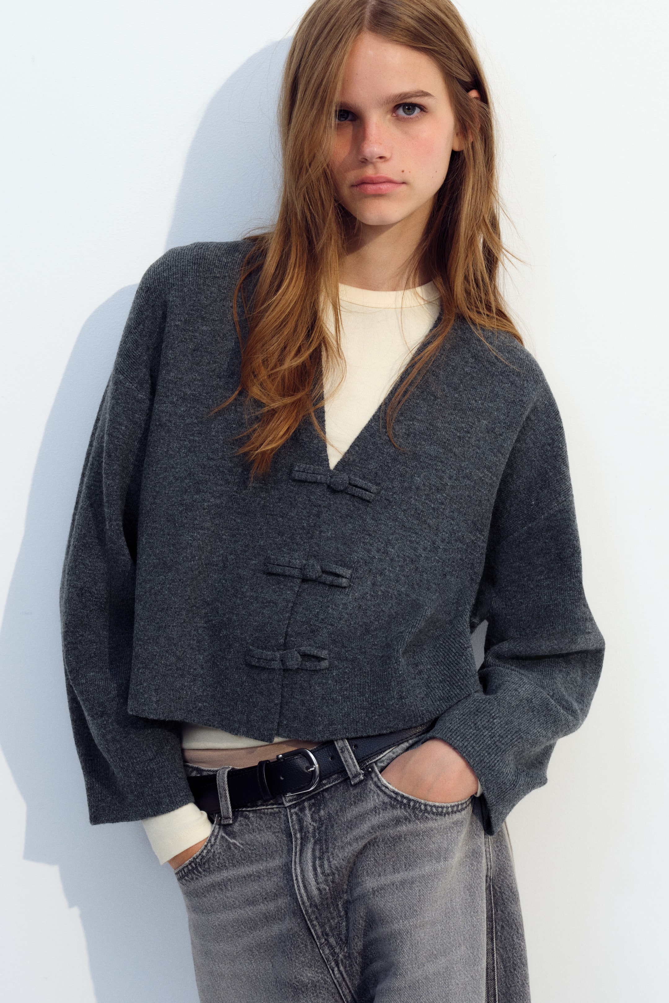 Ampliar la imagen: La modelo lleva un cárdigan tejido gris de corte holgado y corto, con escote en V, botones tipo nudo al frente y mangas largas. El cárdigan se combina con una prenda superior color crema y jeans de mezclilla grises.