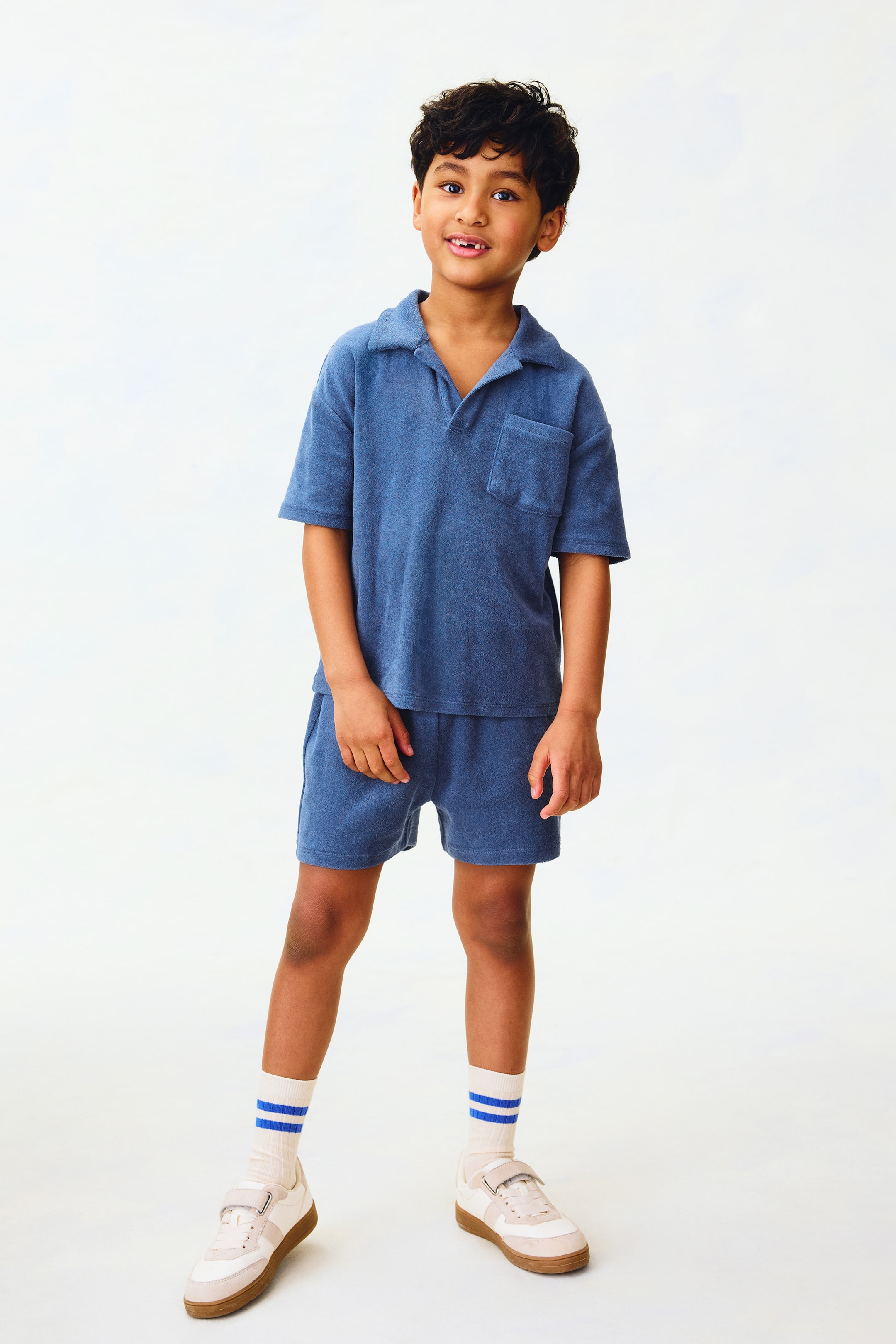 View larger image: A blue toweling polo shirt and matching shorts set is displayed front-facing. The short-sleeved polo shirt features a textured fabric, a spread collar, a V-neck opening, and a left chest patch pocket, while the loose-fitting shorts are mid-thigh length.