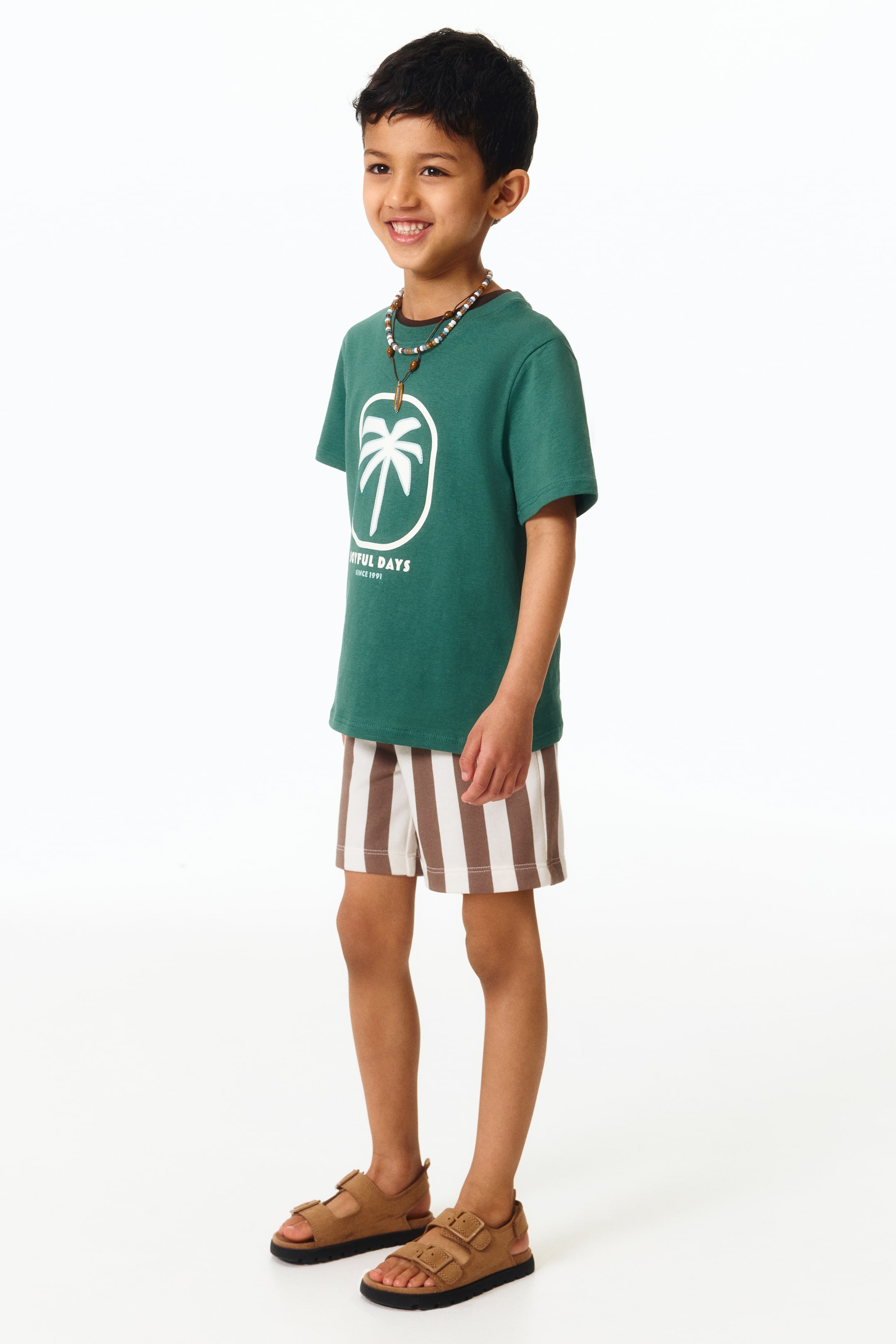 Agrandir l'image: Un t-shirt à manches courtes vert foncé est présenté, arborant un graphique de palmier blanc et du texte sur le devant. Il est assorti à un short à rayures verticales marron et blanc, atteignant le milieu des cuisses.