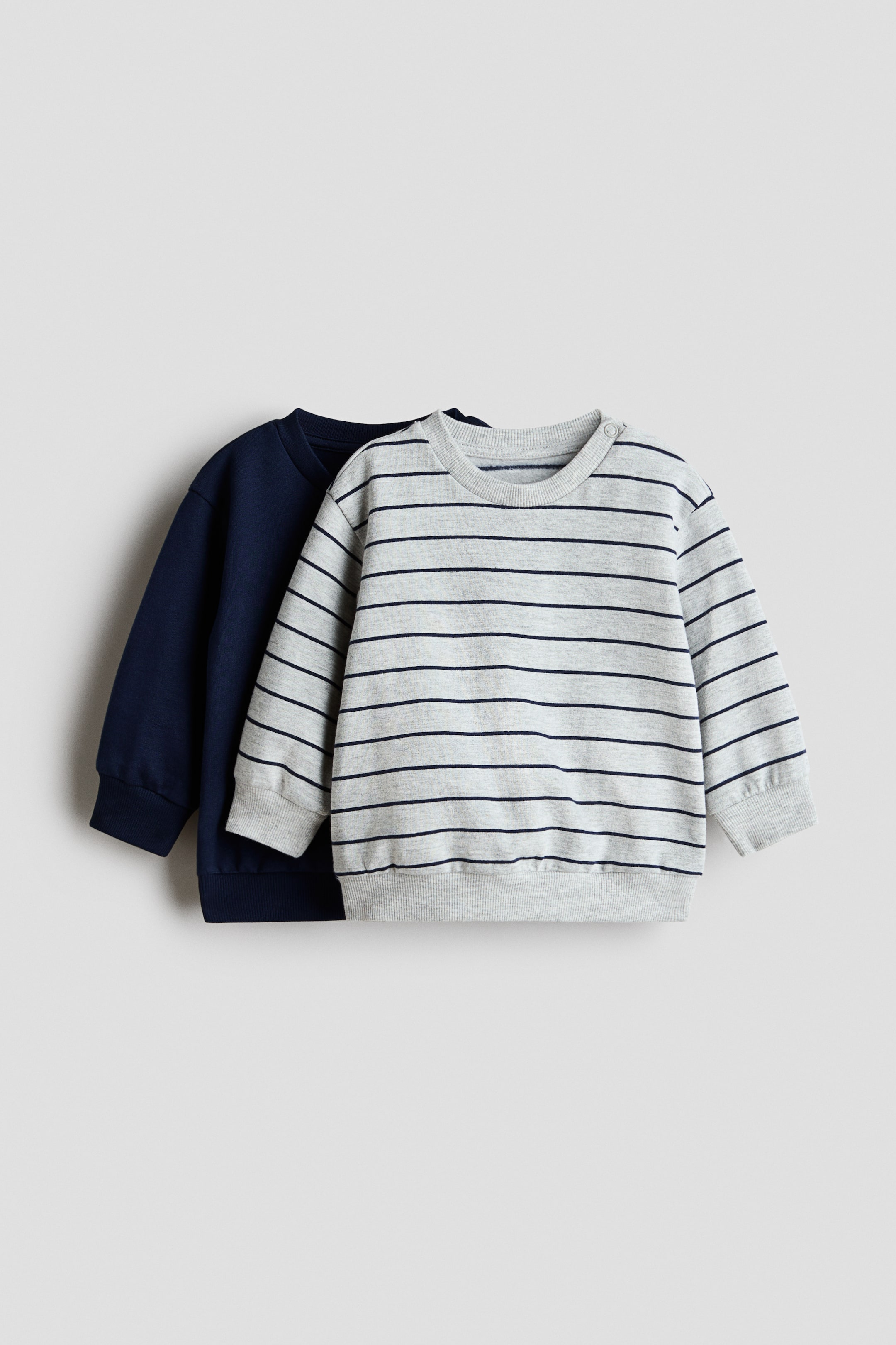 View larger image: Two sweatshirts are displayed, with a light grey striped sweatshirt layered over a solid navy blue one. The grey sweatshirt showcases horizontal navy blue stripes, two snap fasteners on the left shoulder, long sleeves, and ribbed trim at the neck, cuffs, and hem. The navy blue sweatshirt also features long sleeves and ribbed cuffs and hem.