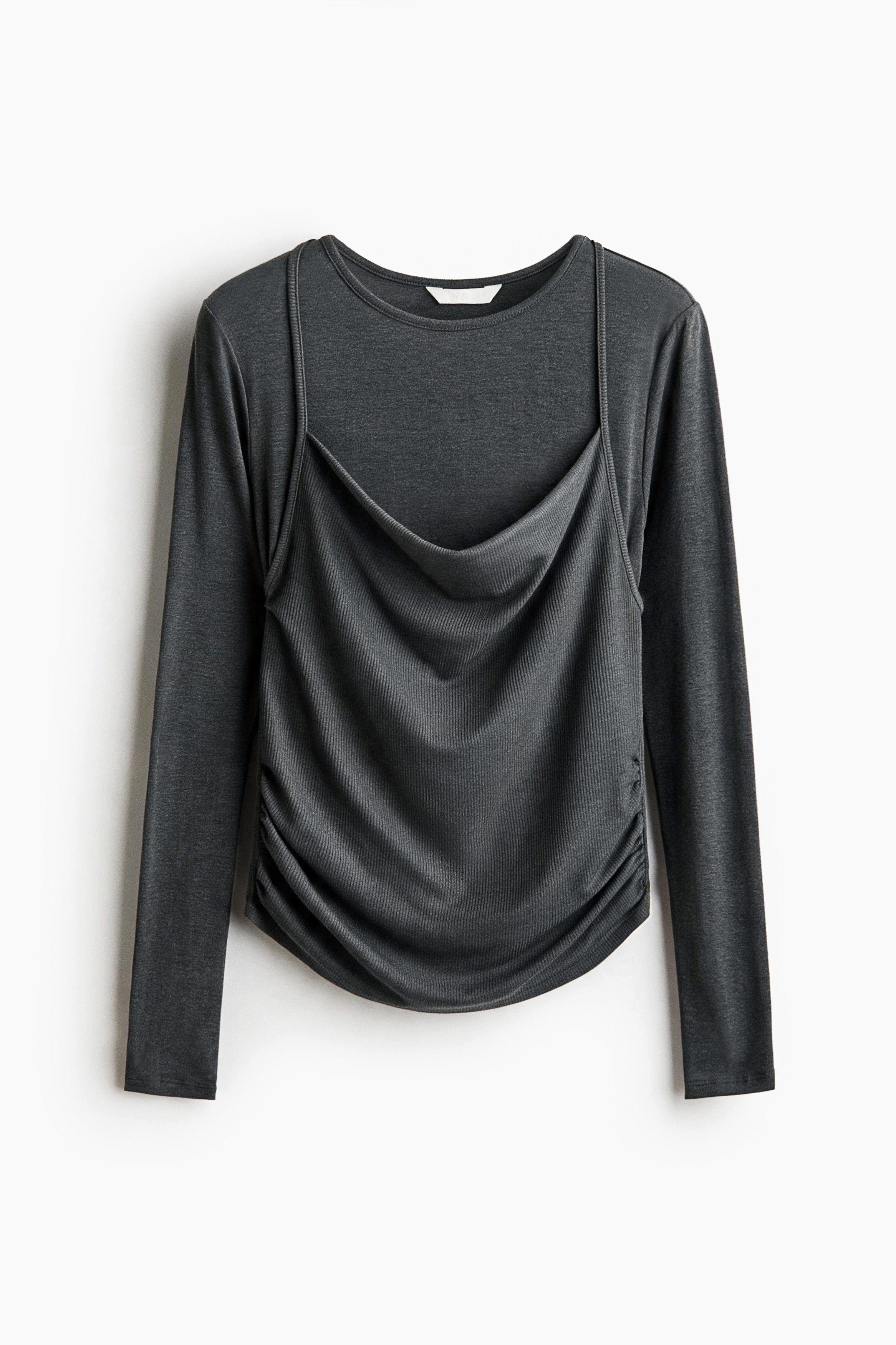 The model is wearing a dark gray, long-sleeved jersey top featuring a round neckline with a draped cowl-neck front panel and thin halterneck straps. The top has gathered side seams at the waist and is styled with loose, wide-leg dark denim trousers.