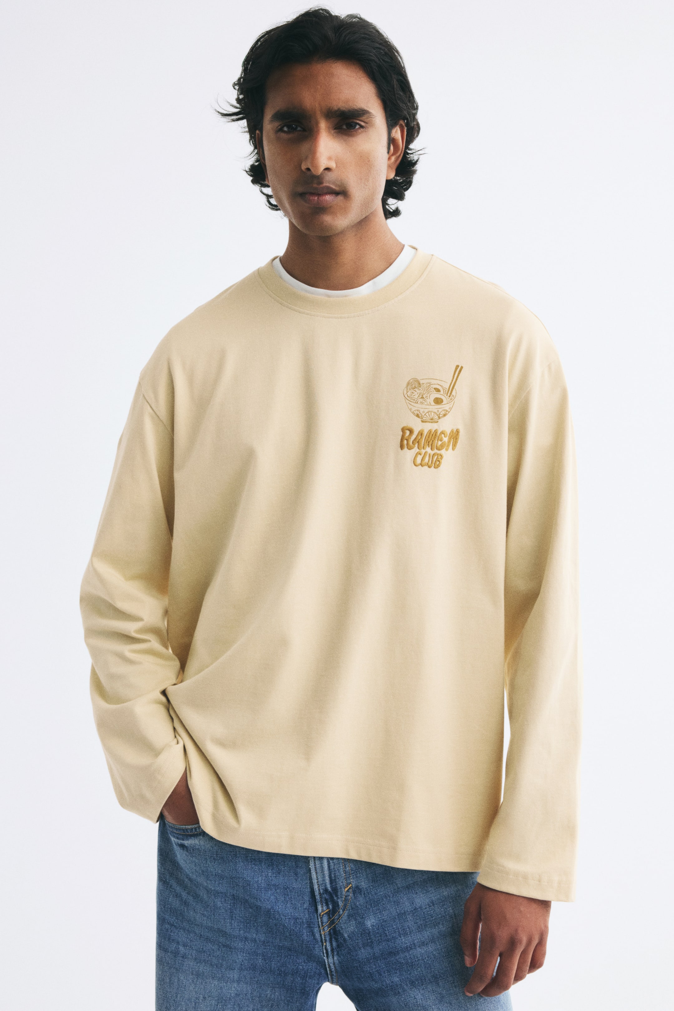 View larger image: A light beige, long-sleeve T-shirt featuring dropped shoulders, a ribbed round neckline, and a graphic of a ramen bowl with the text "RAMEN CLUB" on the chest is being worn by the model. The shirt is layered over a white collar and paired with blue jeans, with the model keeping one hand in a front pocket.